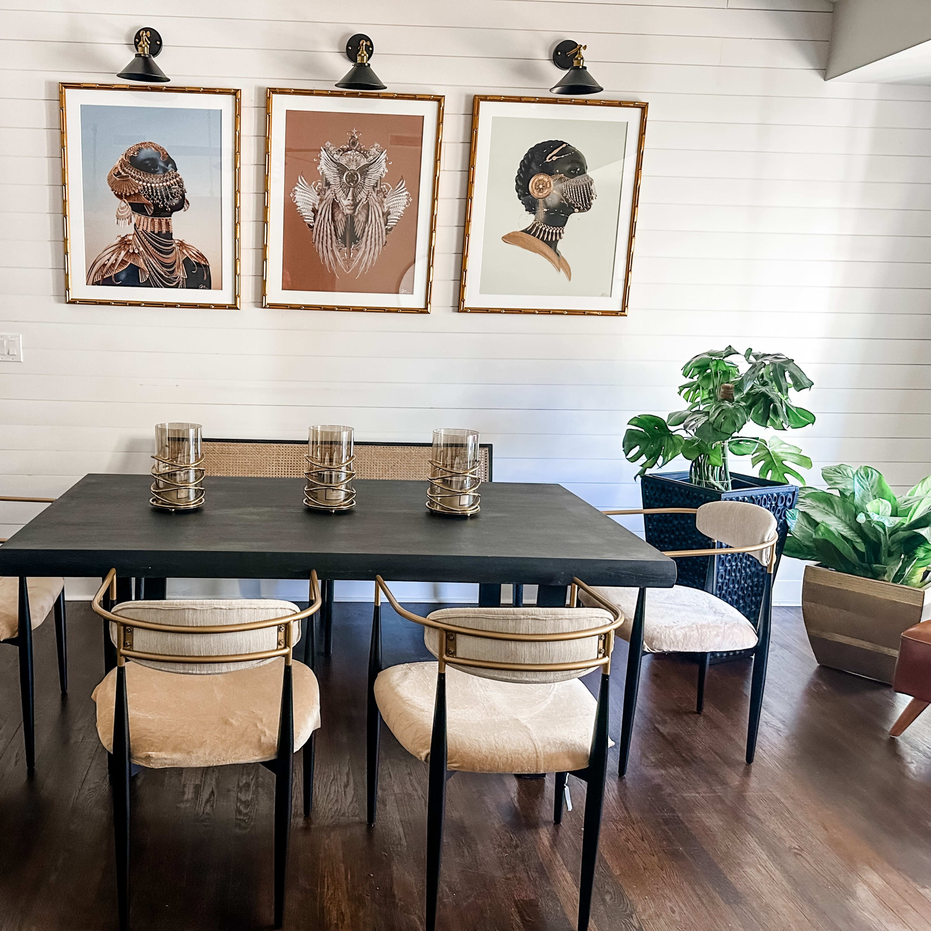 A dining area features a black table surrounded by six chairs, with three framed artworks hung on the white wall and a door to the left.