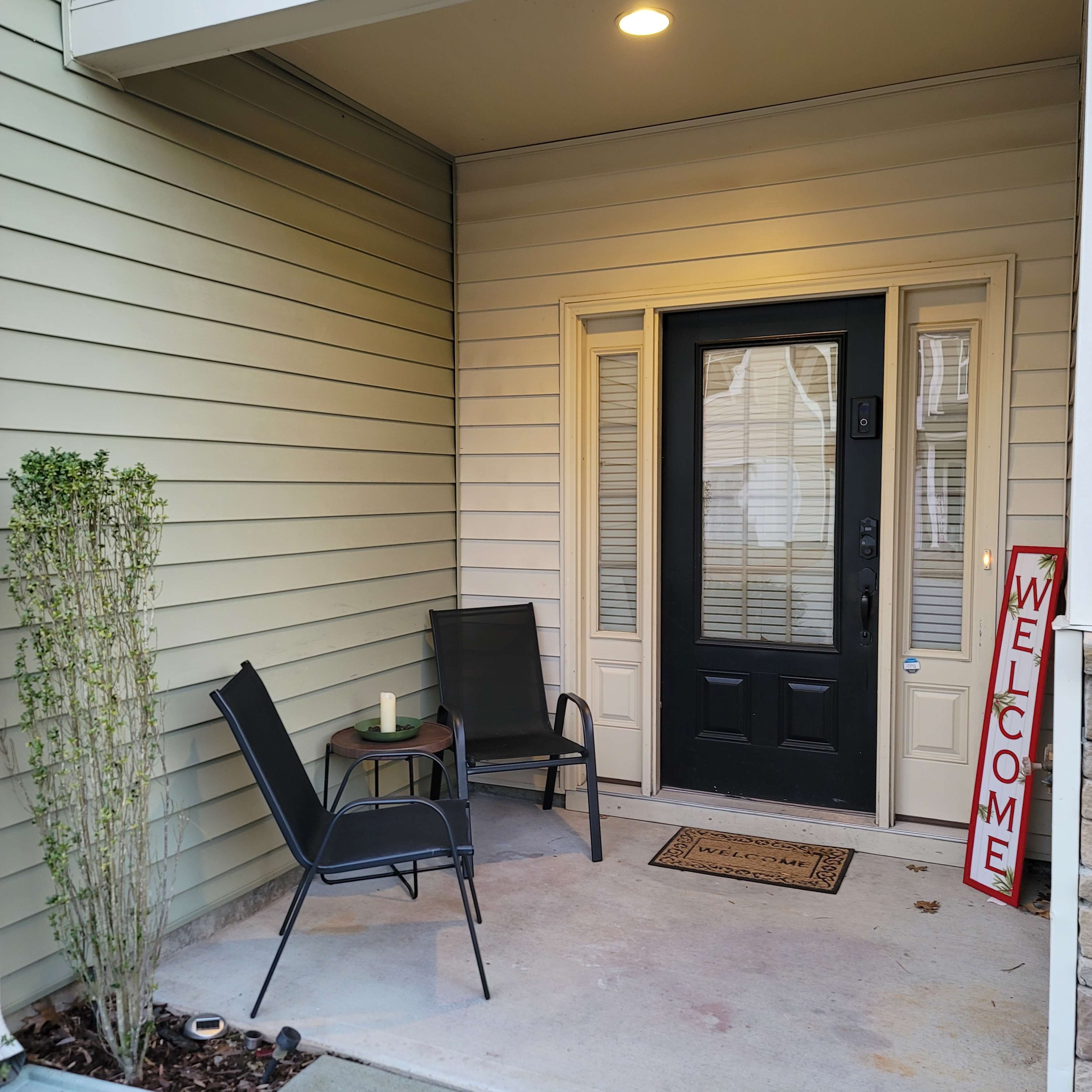 A front porch with two chairs and a small table beside a black door, featuring a "Welcome" sign.