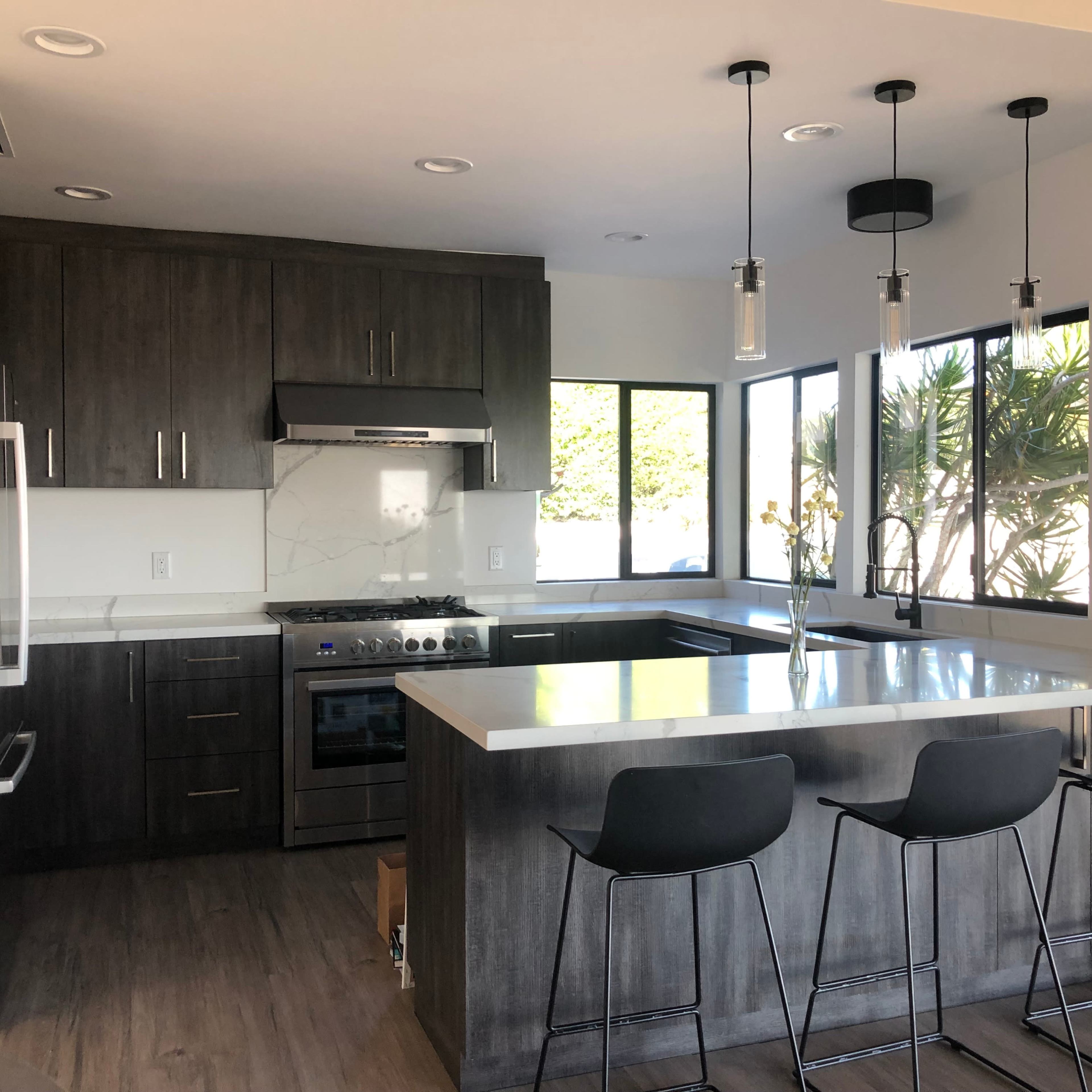 A modern kitchen with dark cabinetry, stainless steel appliances, and a bar with three stools facing a window.