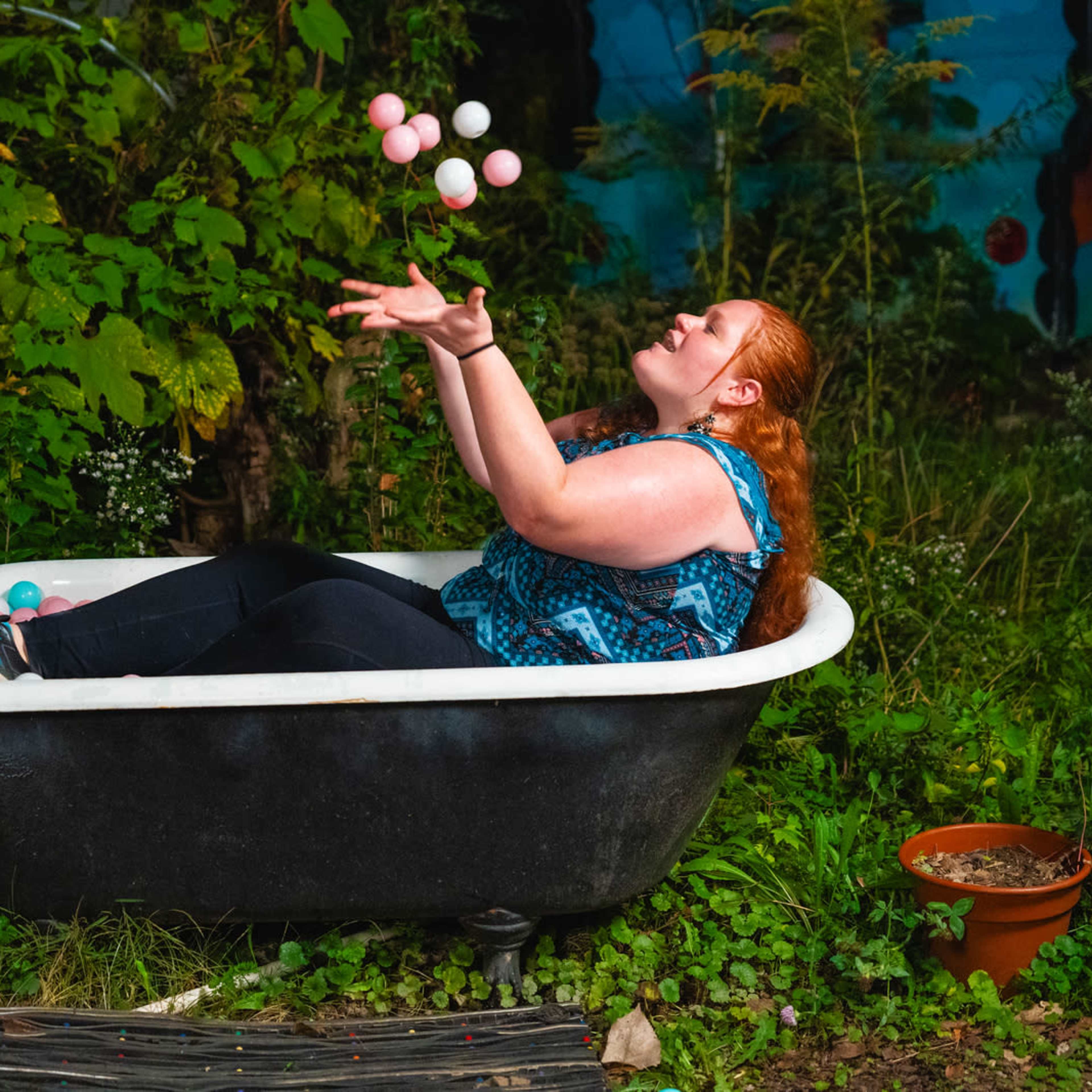 A woman with curly red hair is juggling colorful balls while sitting in a vintage bathtub surrounded by greenery and plants.
