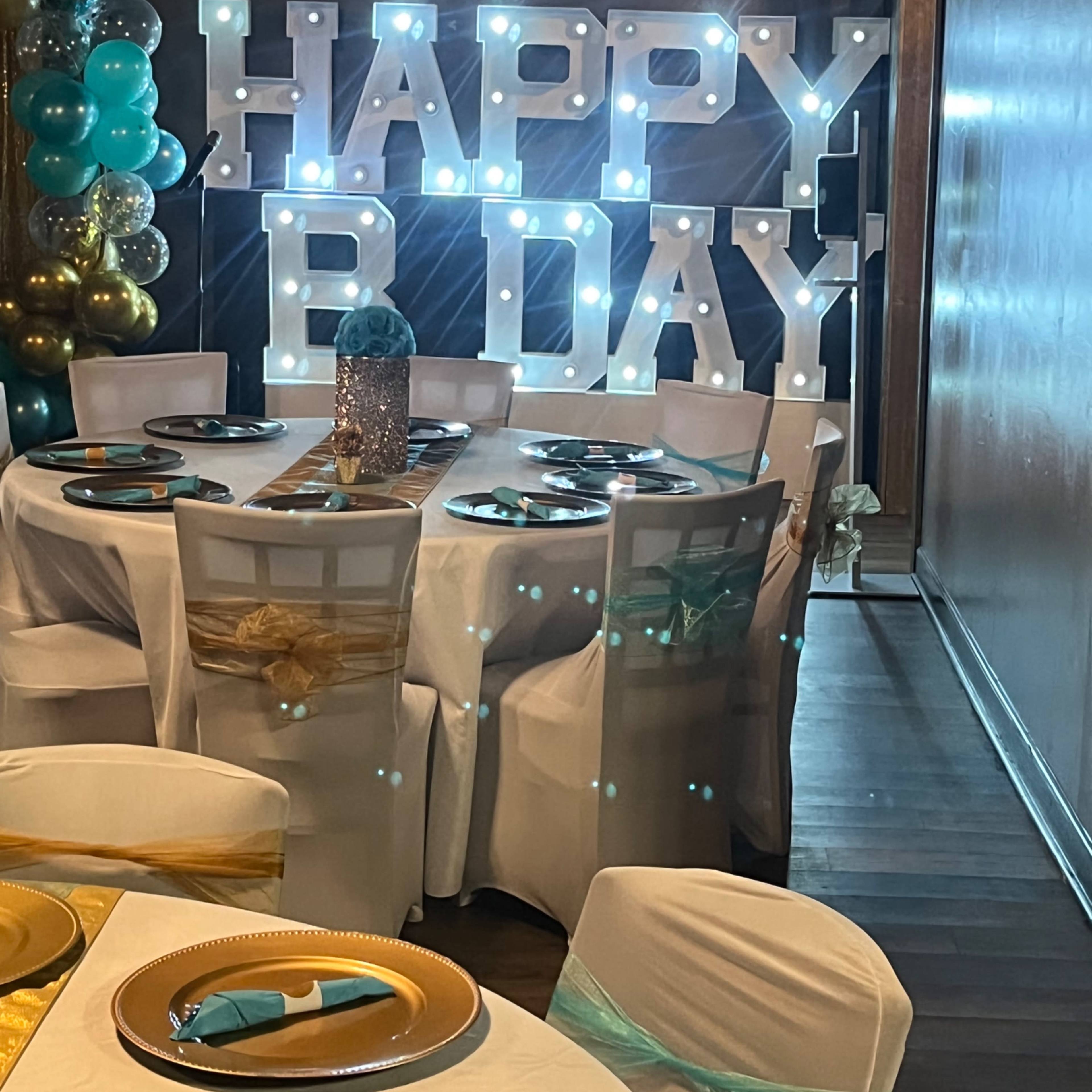 A festive birthday celebration set up with a "HAPPY B DAY" sign illuminated by lights, decorated tables, and balloons in the background.