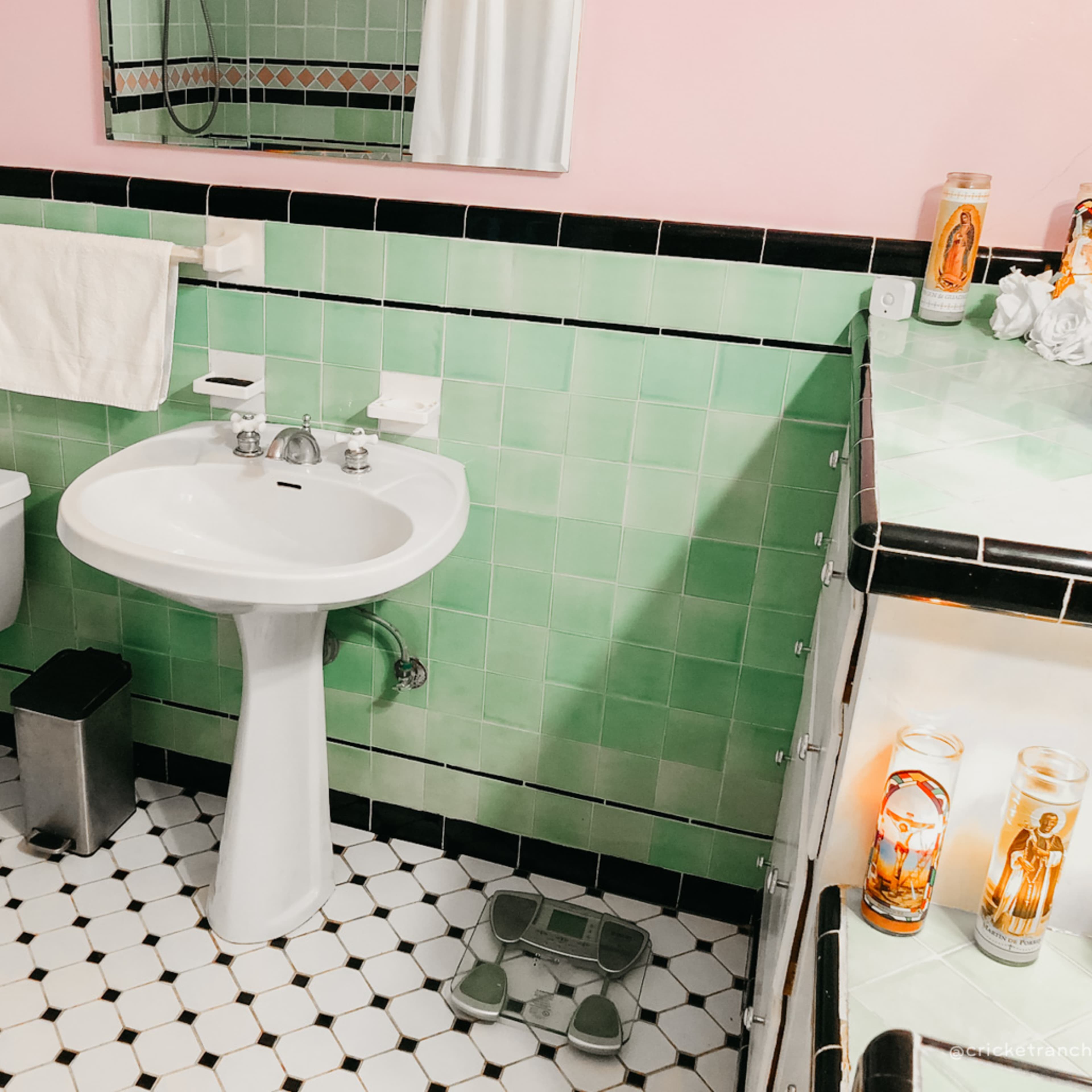 A small bathroom with mint green tiles, a white sink and toilet, a scale, and a row of candles on the countertop.