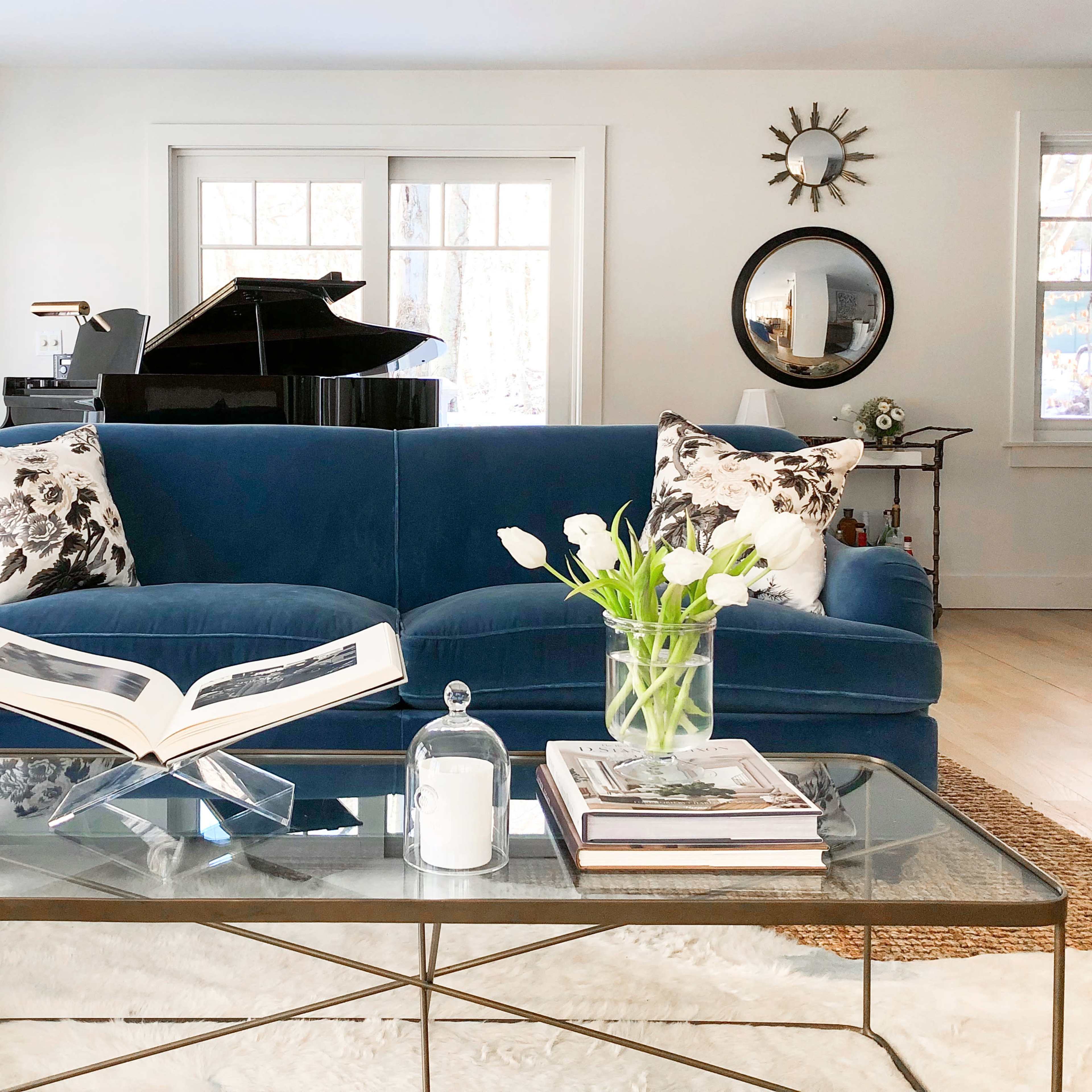 A blue sofa with floral-patterned pillows is positioned in a bright room, featuring a glass coffee table and a grand piano in the background.