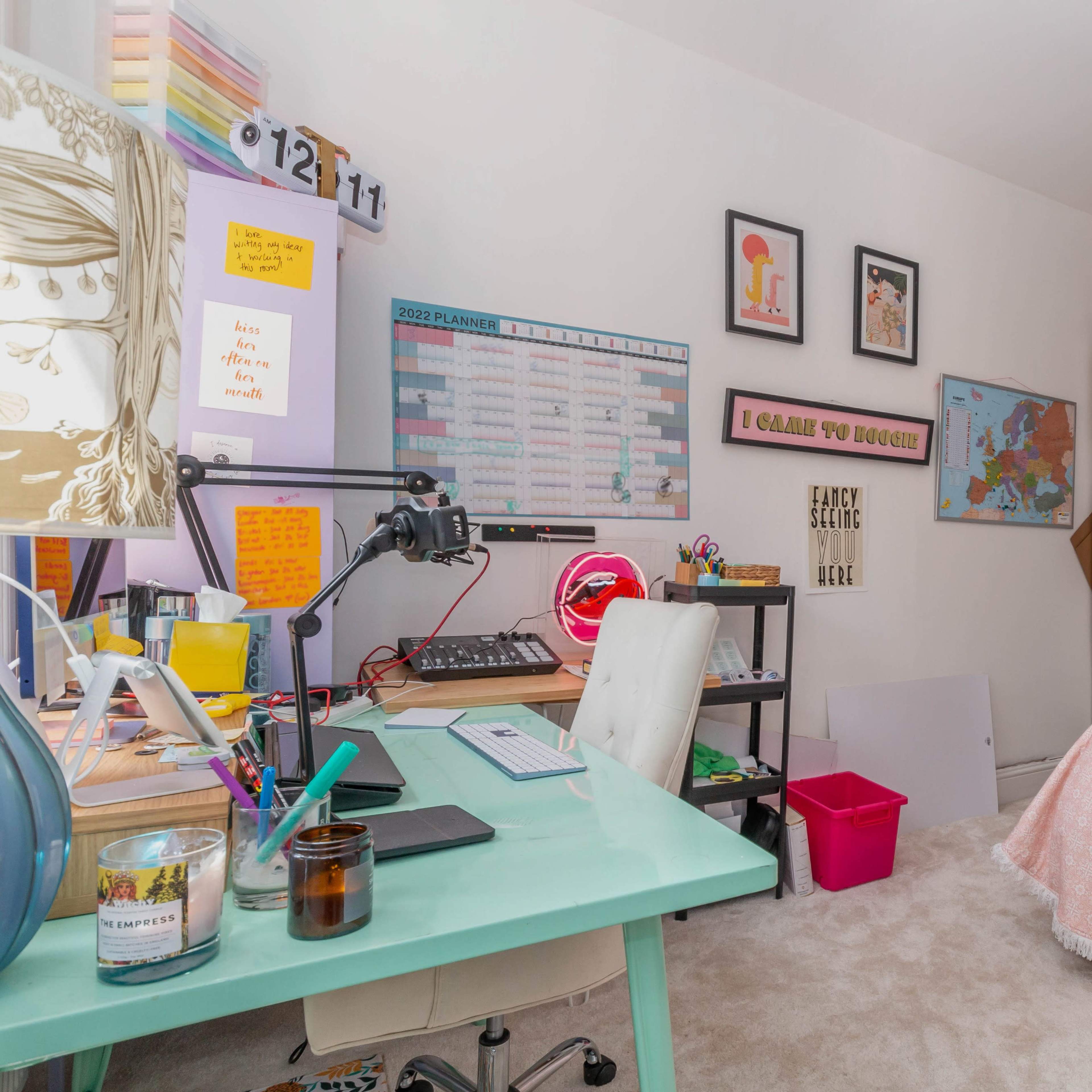 A small bedroom is organized into a workspace with a desk, computer, and decorative elements, alongside a bed covered with a pink throw and a bookshelf.