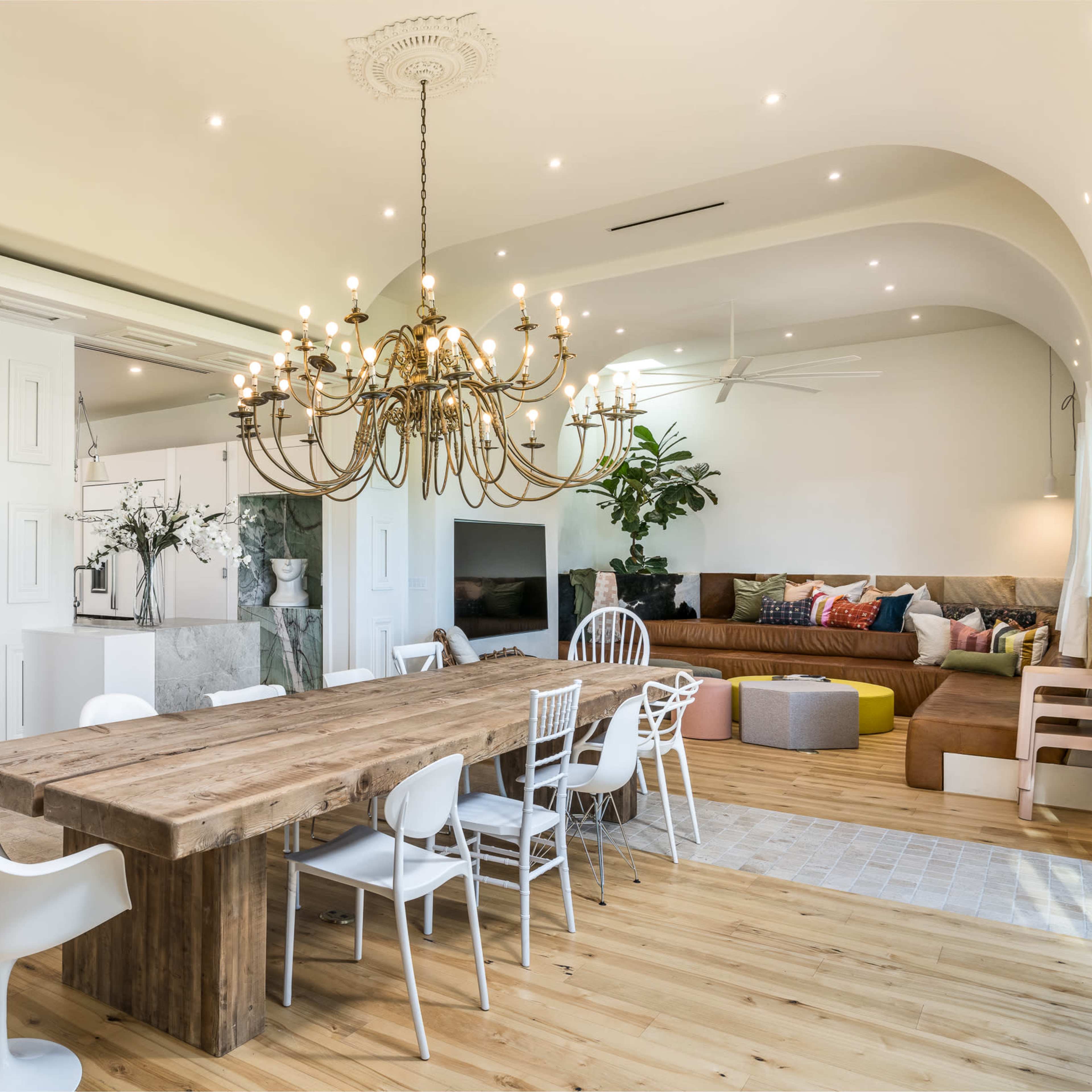 A modern dining area with a large wooden table surrounded by white chairs, a chandelier overhead, and a spacious living area in the background with a sectional sofa.