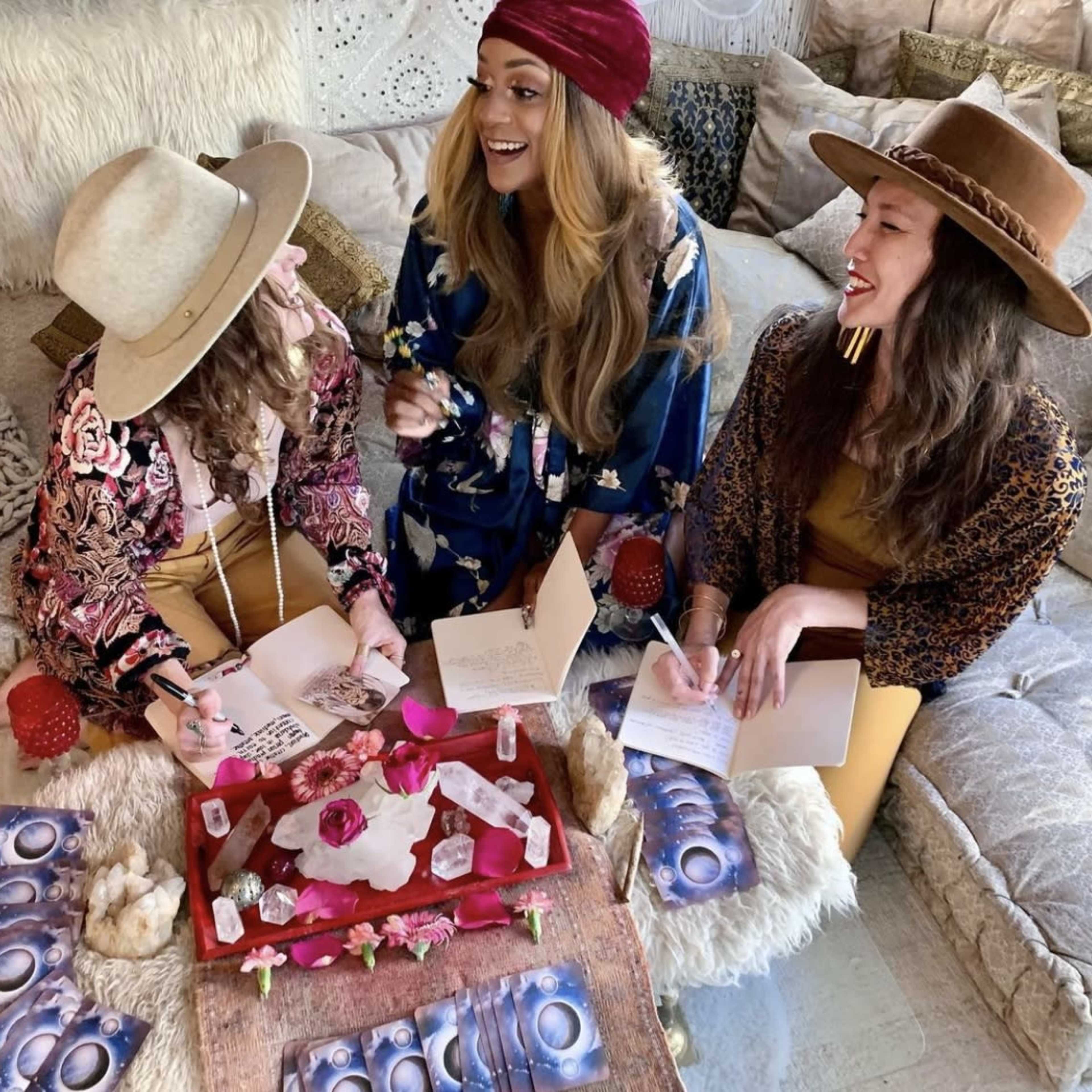Three women wearing hats sit around a table covered with crystals and a rose, writing in journals and sharing laughter.