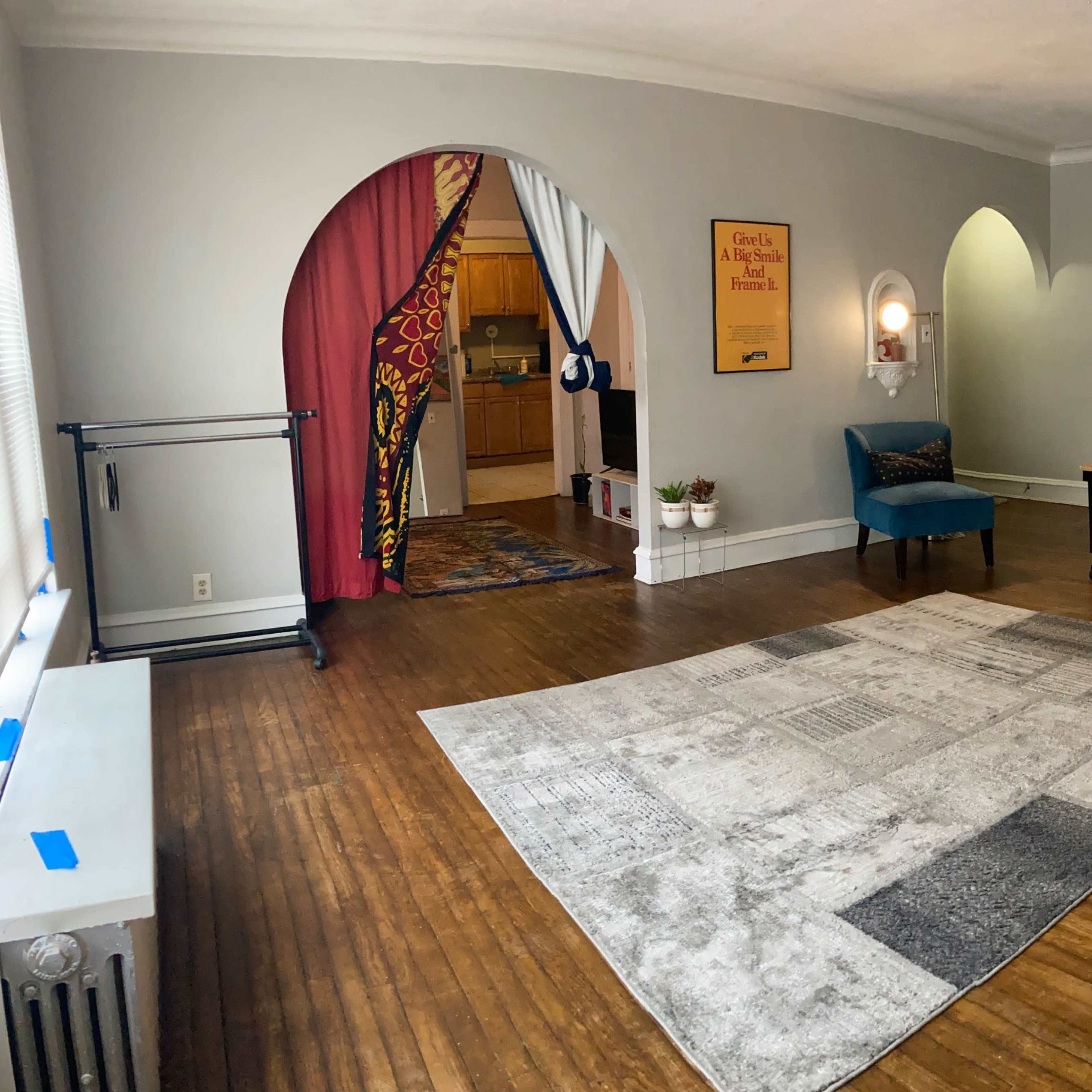 A spacious room with a wooden floor, a large area rug, a beige couch, a table with a chair, and an archway leading to another area with a kitchenette.