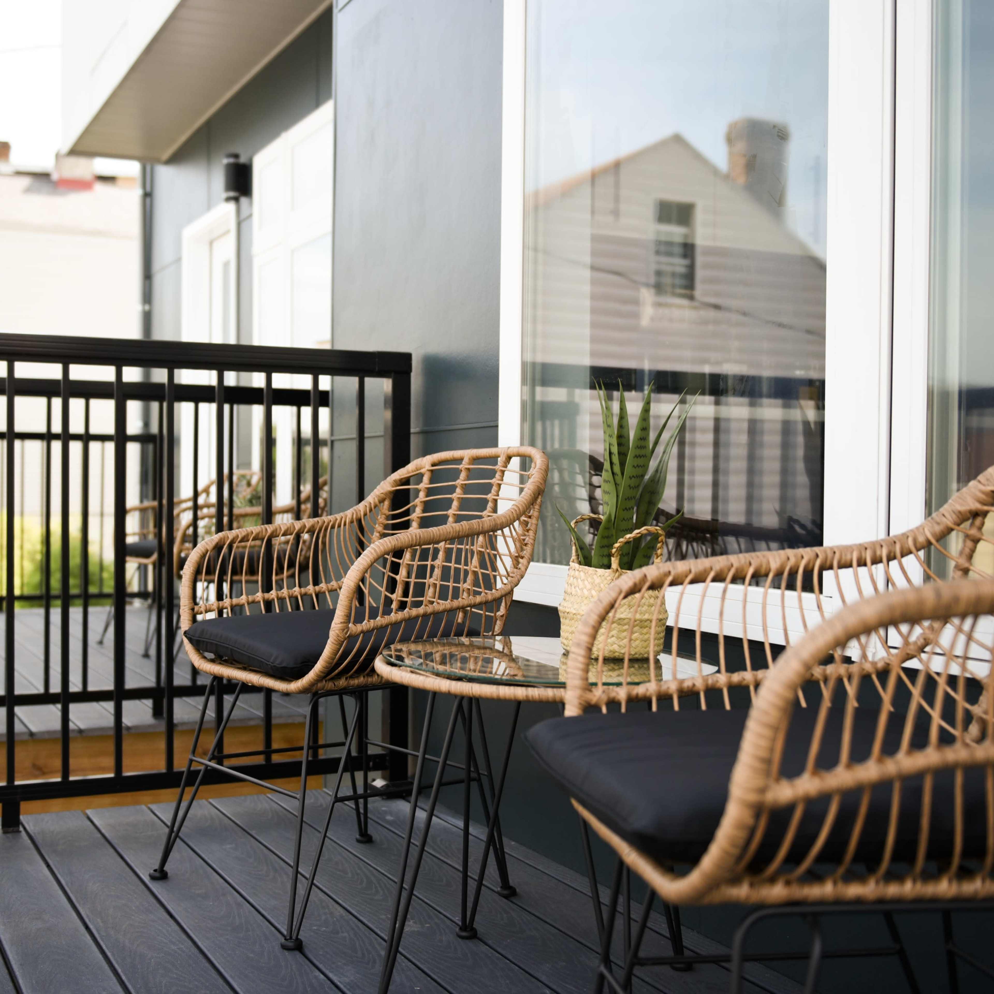 A balcony featuring two wicker chairs and a small table, with a railing and plants visible in the background.