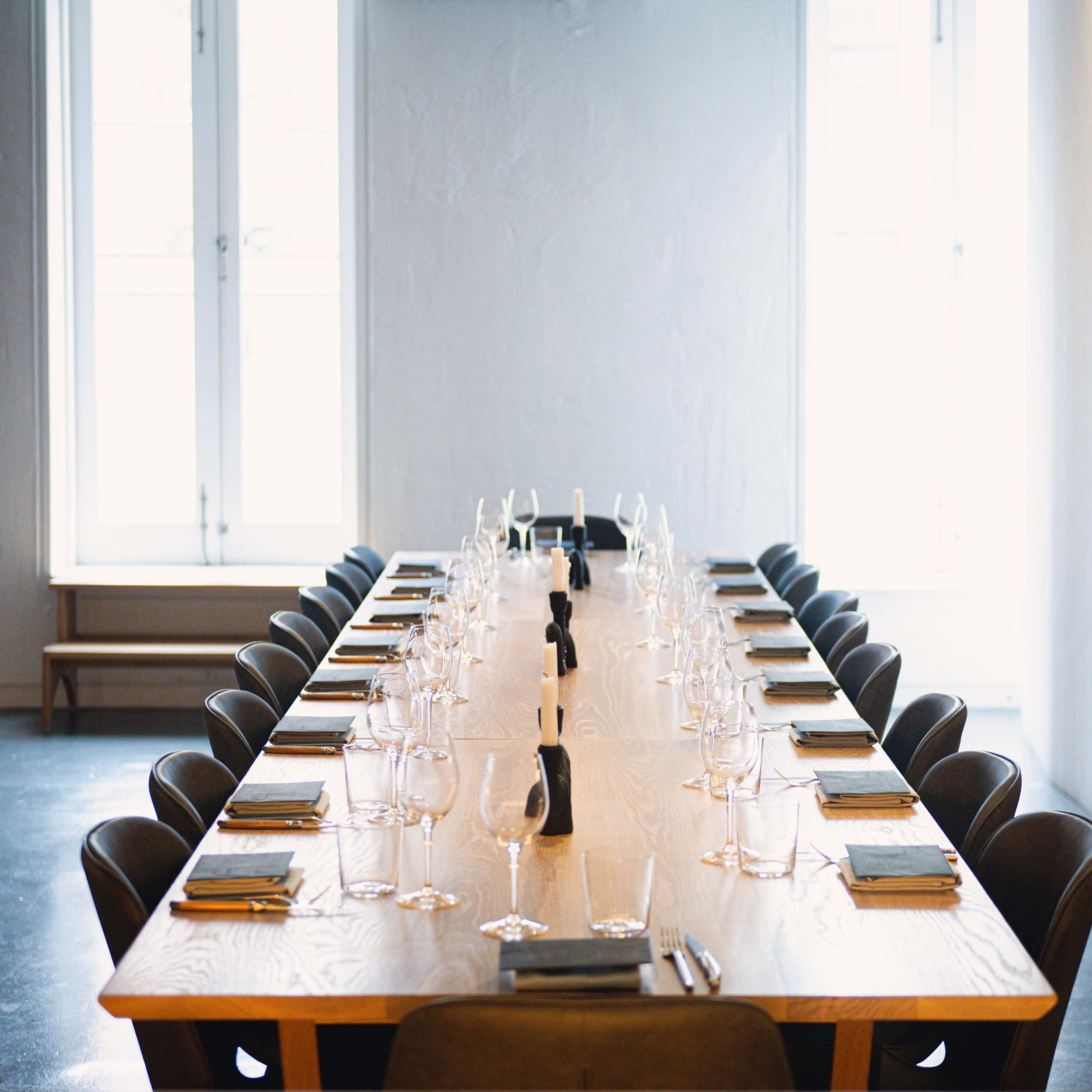 A long wooden dining table is set with plates, utensils, and glasses, positioned under large windows in a minimalist interior space.