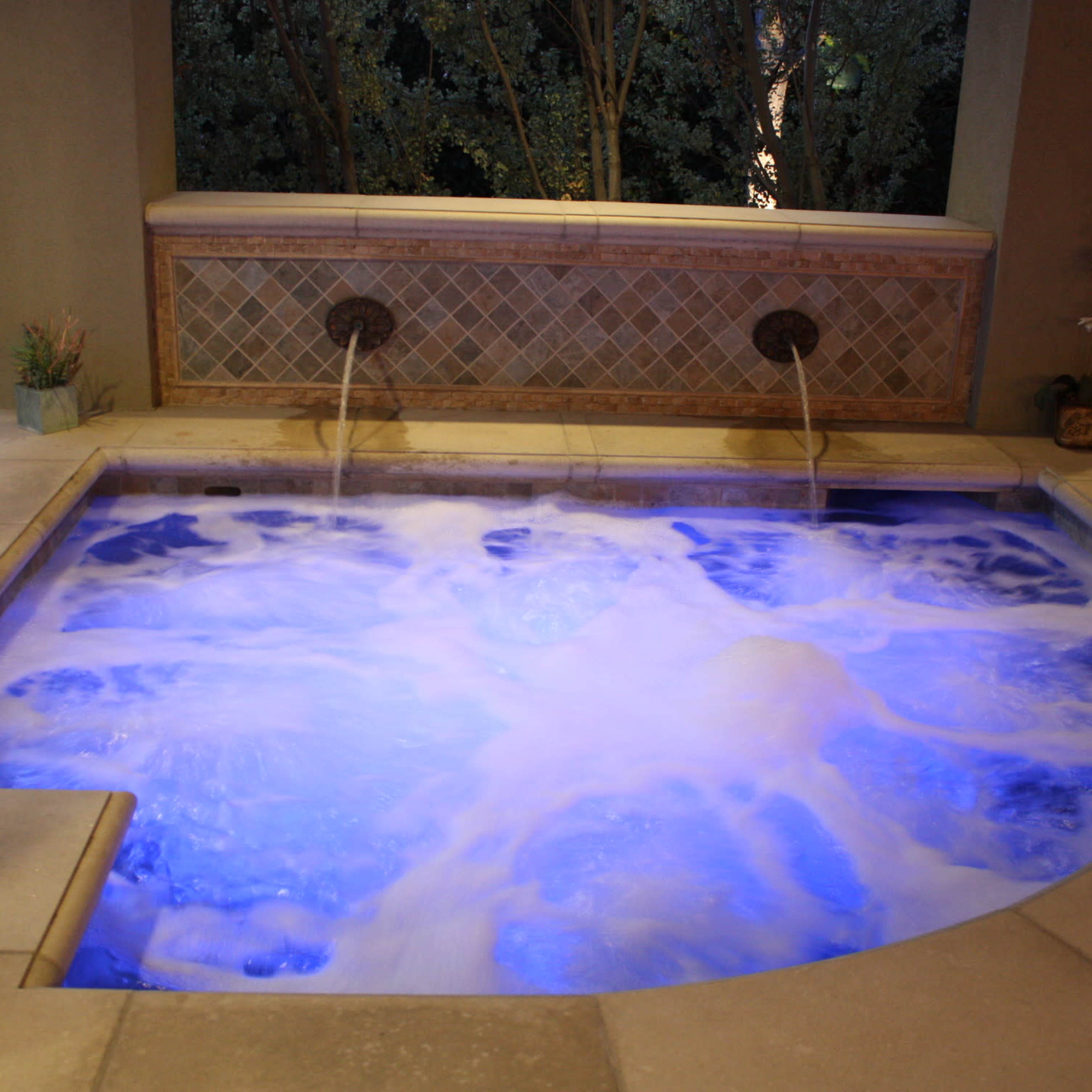 A hot tub with bubbling water and illuminated with blue lighting, set against a backdrop of greenery.