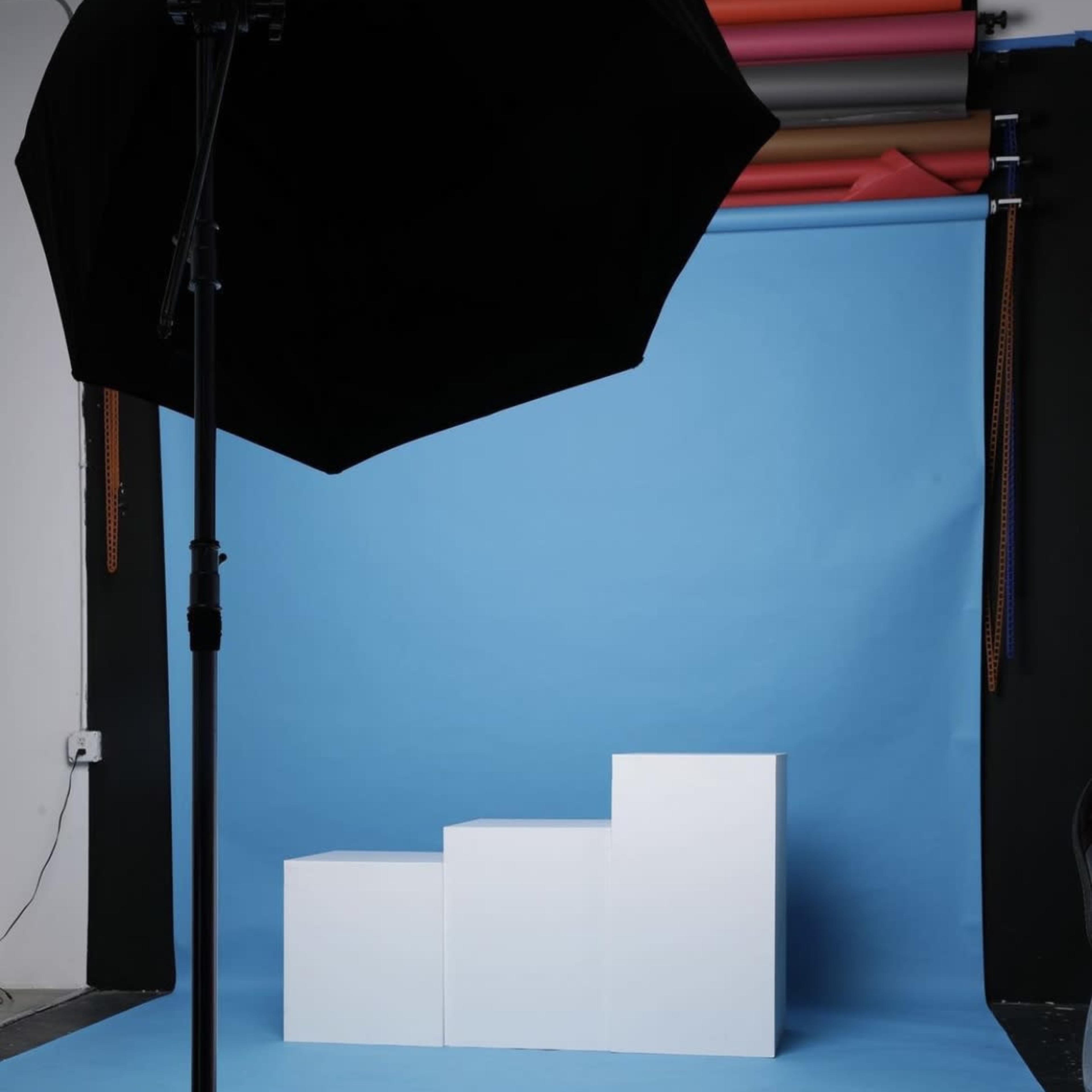 A photography studio setup with a blue backdrop, three white blocks arranged in a staircase formation, and a large black umbrella reflector.