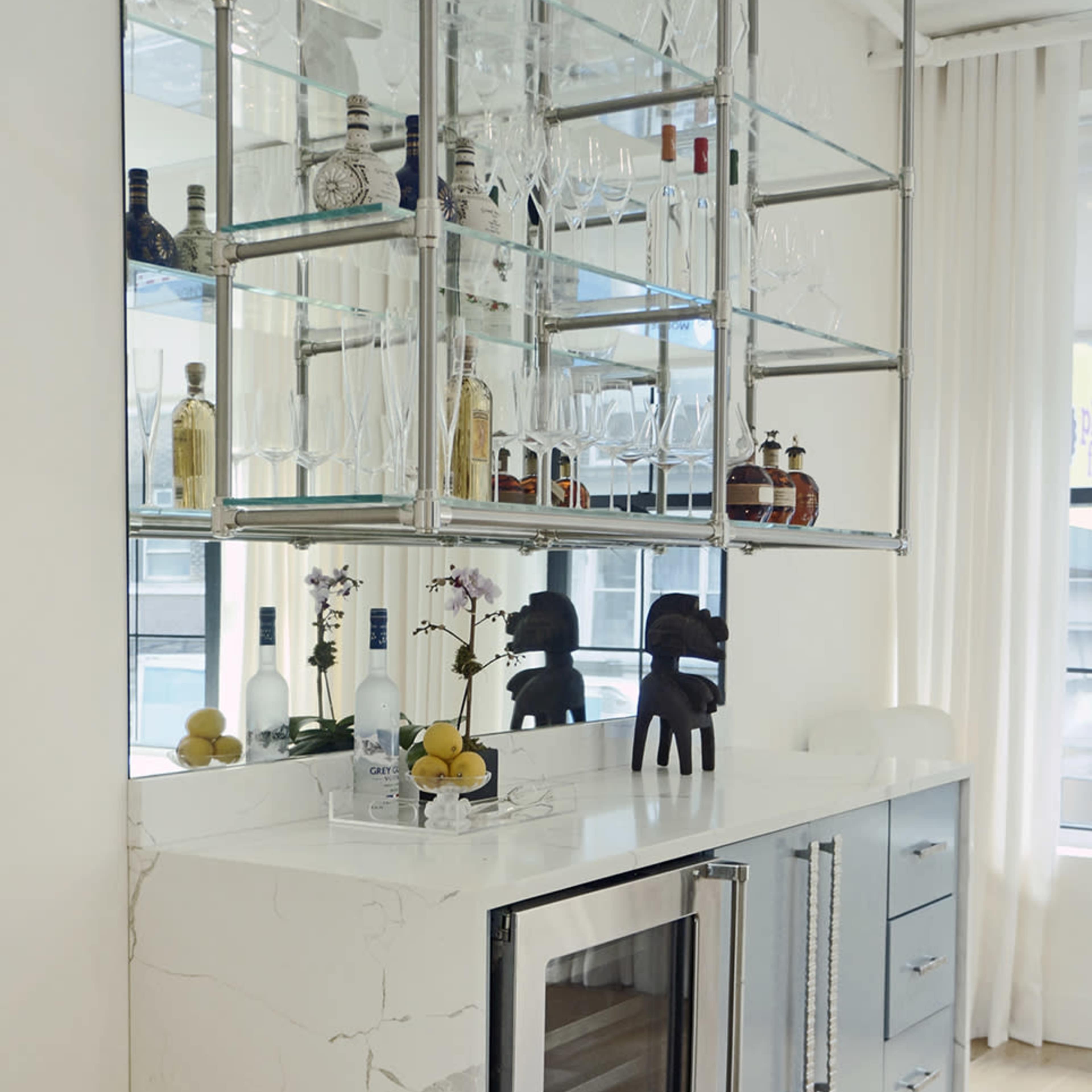 A modern kitchen bar area features a sleek countertop with a built-in wine cooler, multiple shelves displaying glassware and bottles, and a minimalist design.