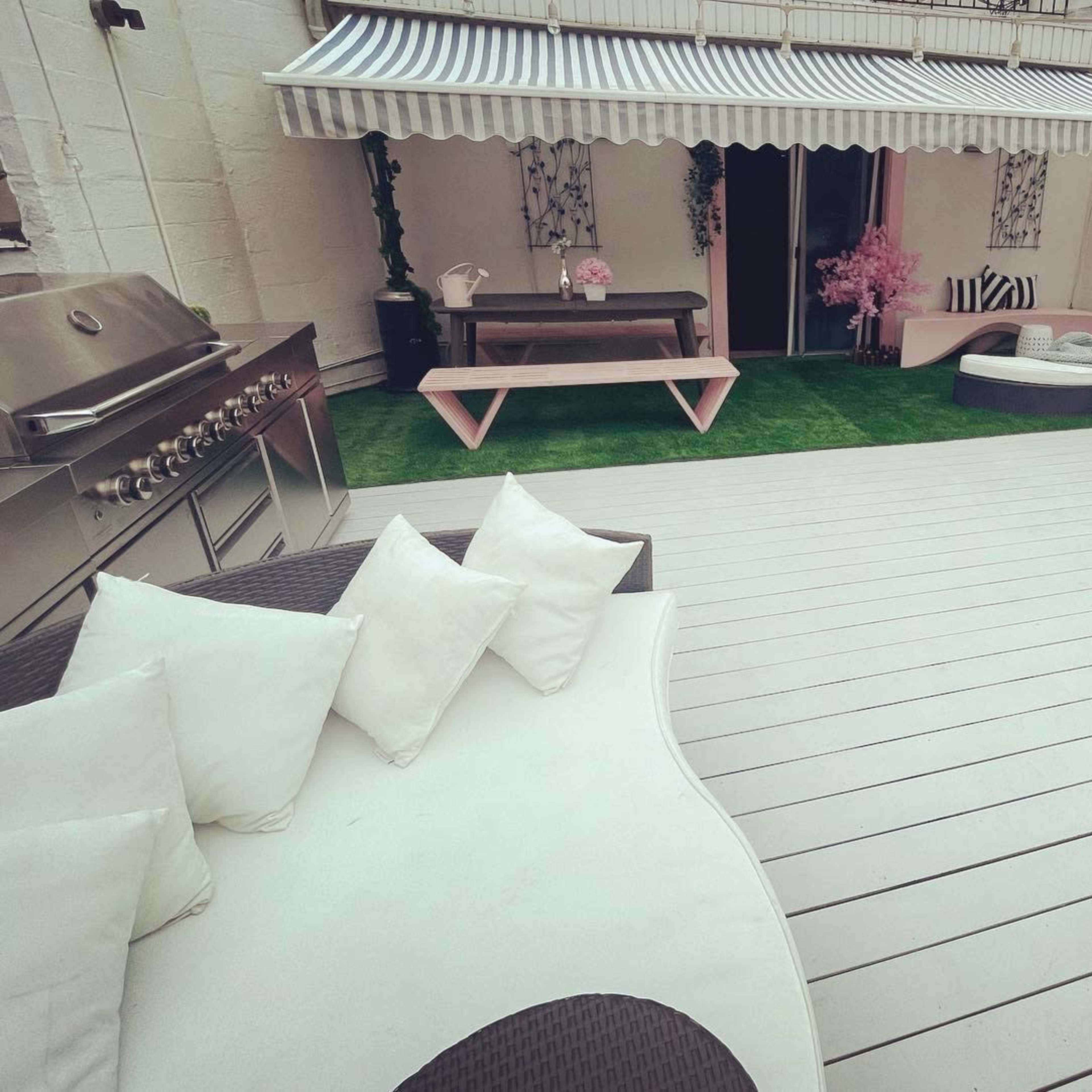 The outdoor space features a gray deck with a grill, a pink table, a daybed, and potted plants under an awning.