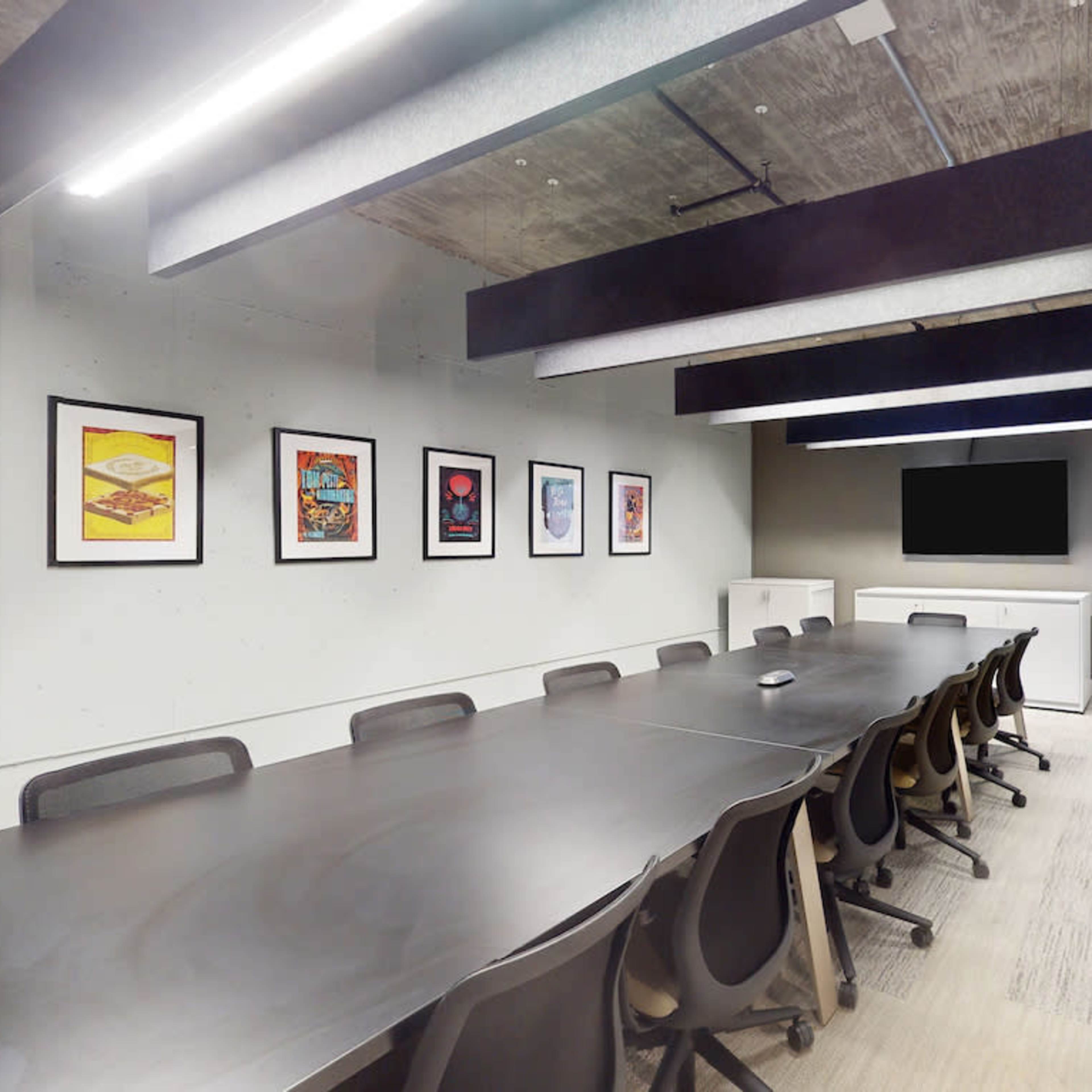 A modern conference room features a large table surrounded by chairs, adorned with framed artwork on the walls and equipped with a TV and whiteboard.