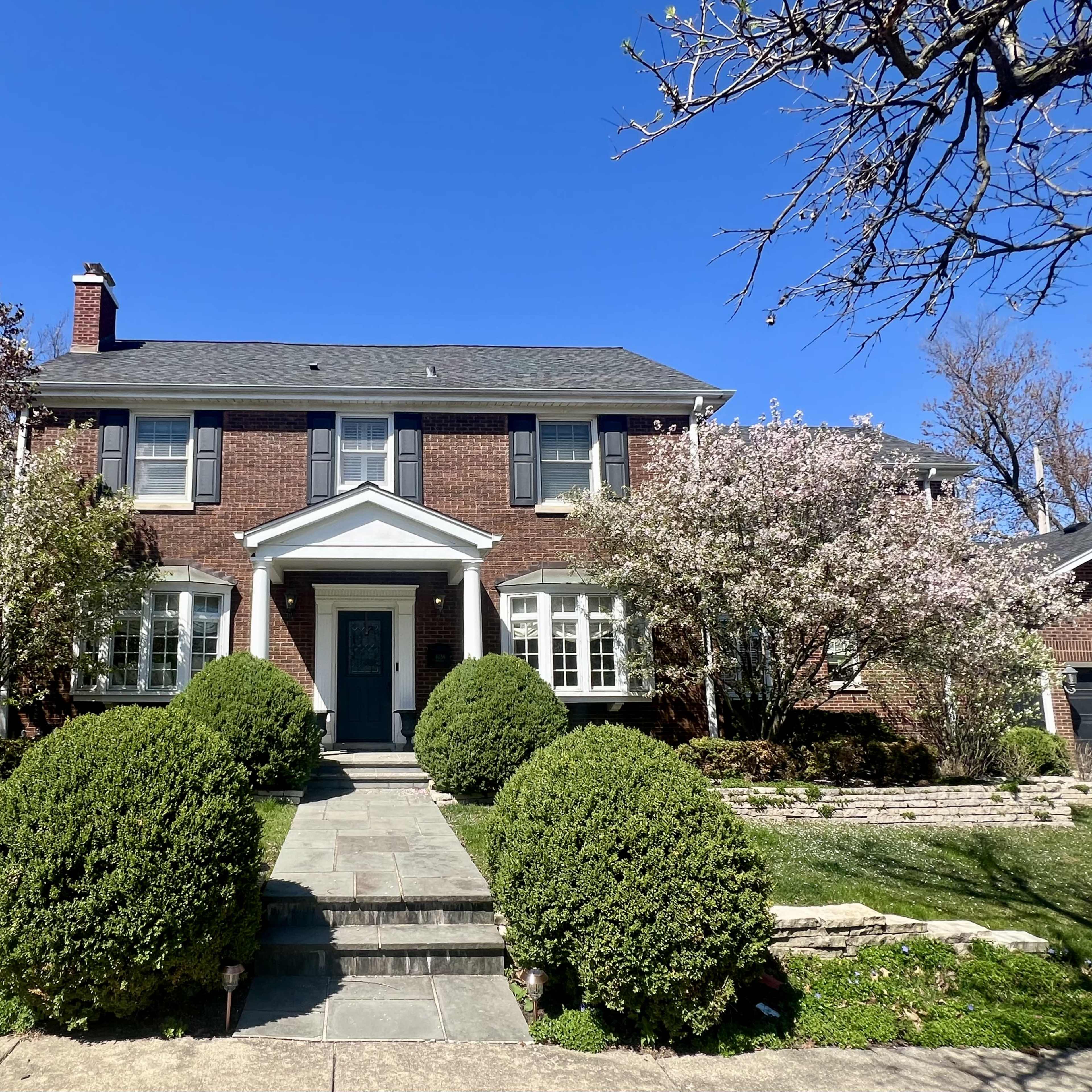 A two-story brick house with large white-framed windows, flanked by flowering trees and neatly trimmed bushes.