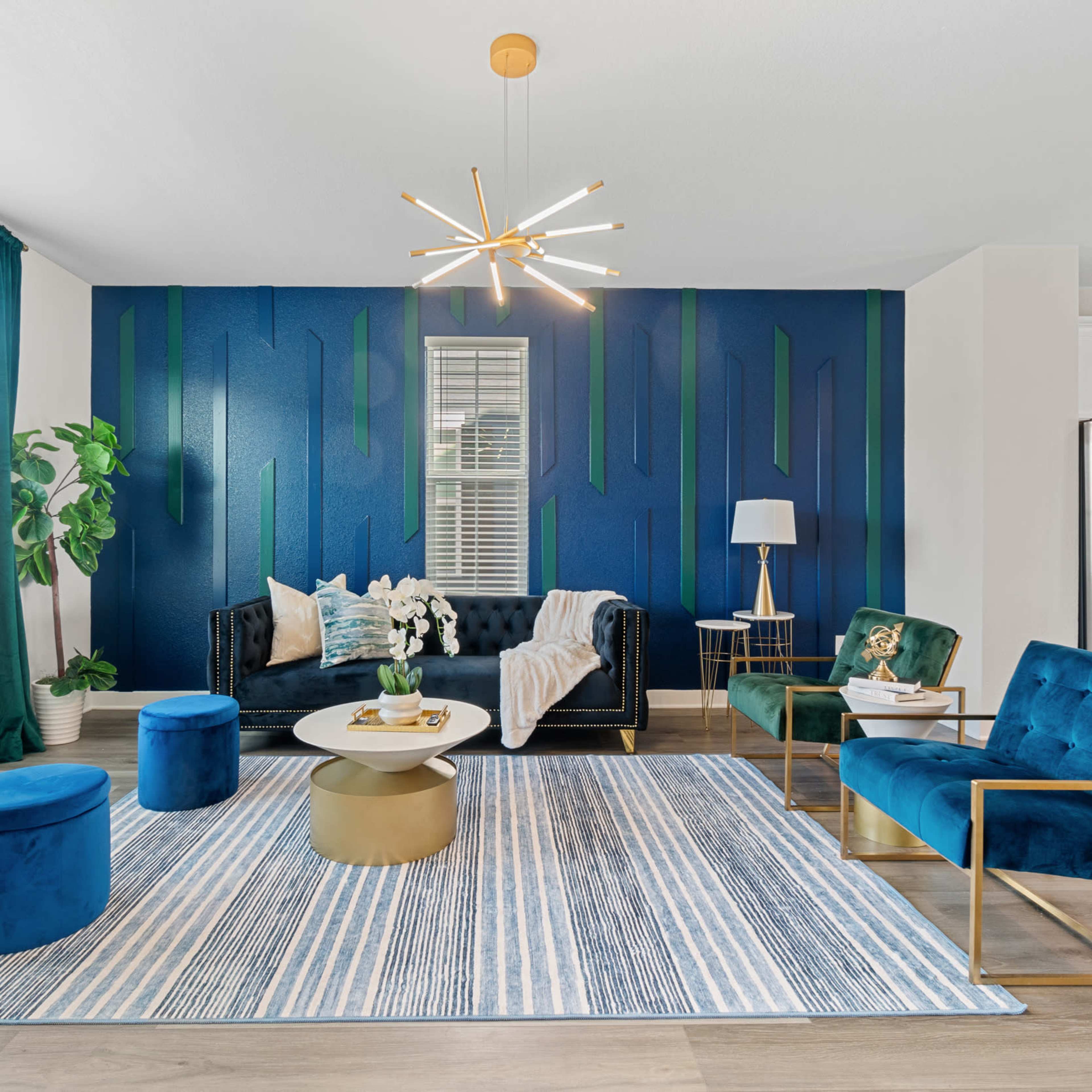 A modern living room with a dark blue accent wall, a black sofa, teal armchairs, a round coffee table, and a striped rug.