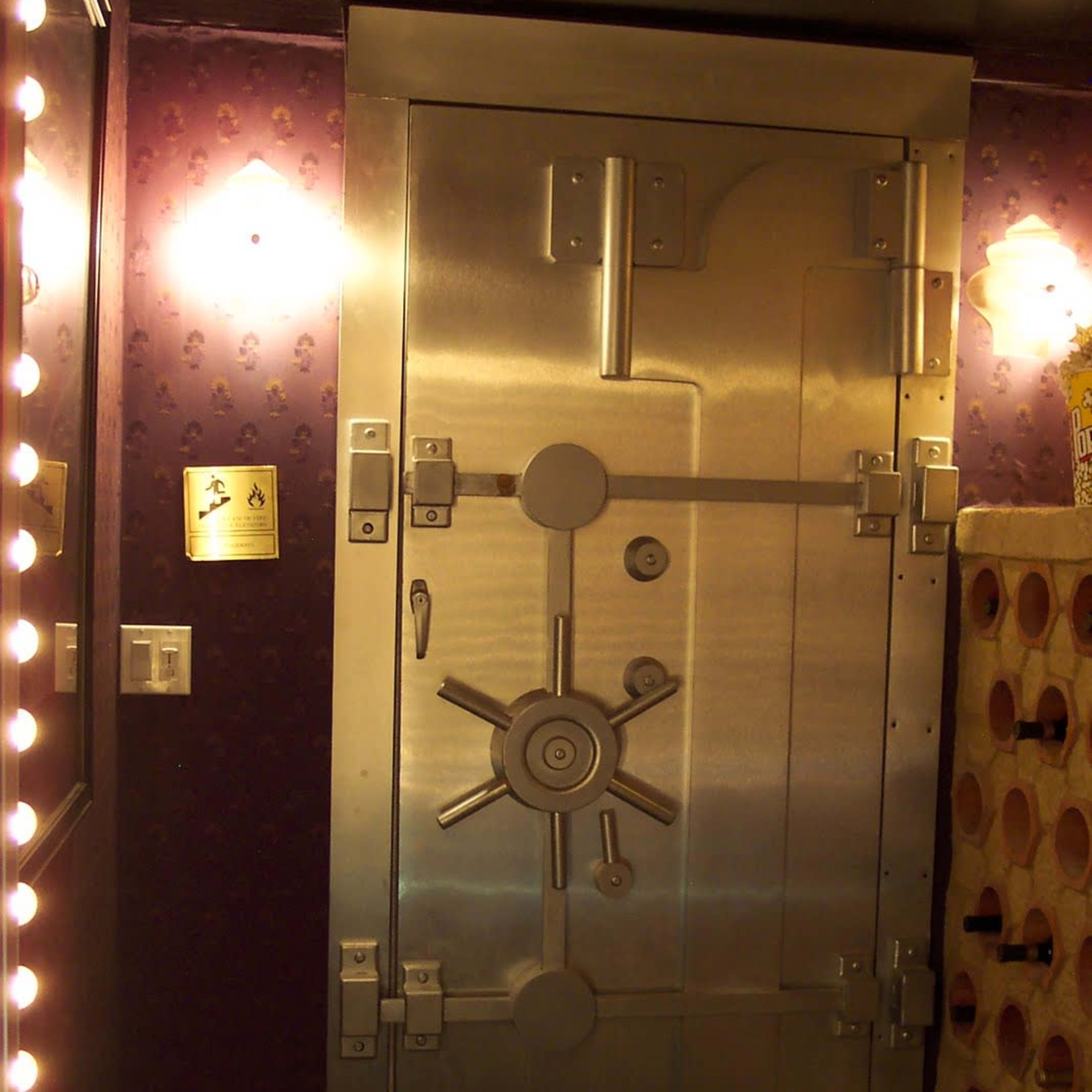 A large, metallic vault door with a wheel mechanism, set against a wall featuring purple wallpaper and bright lights.