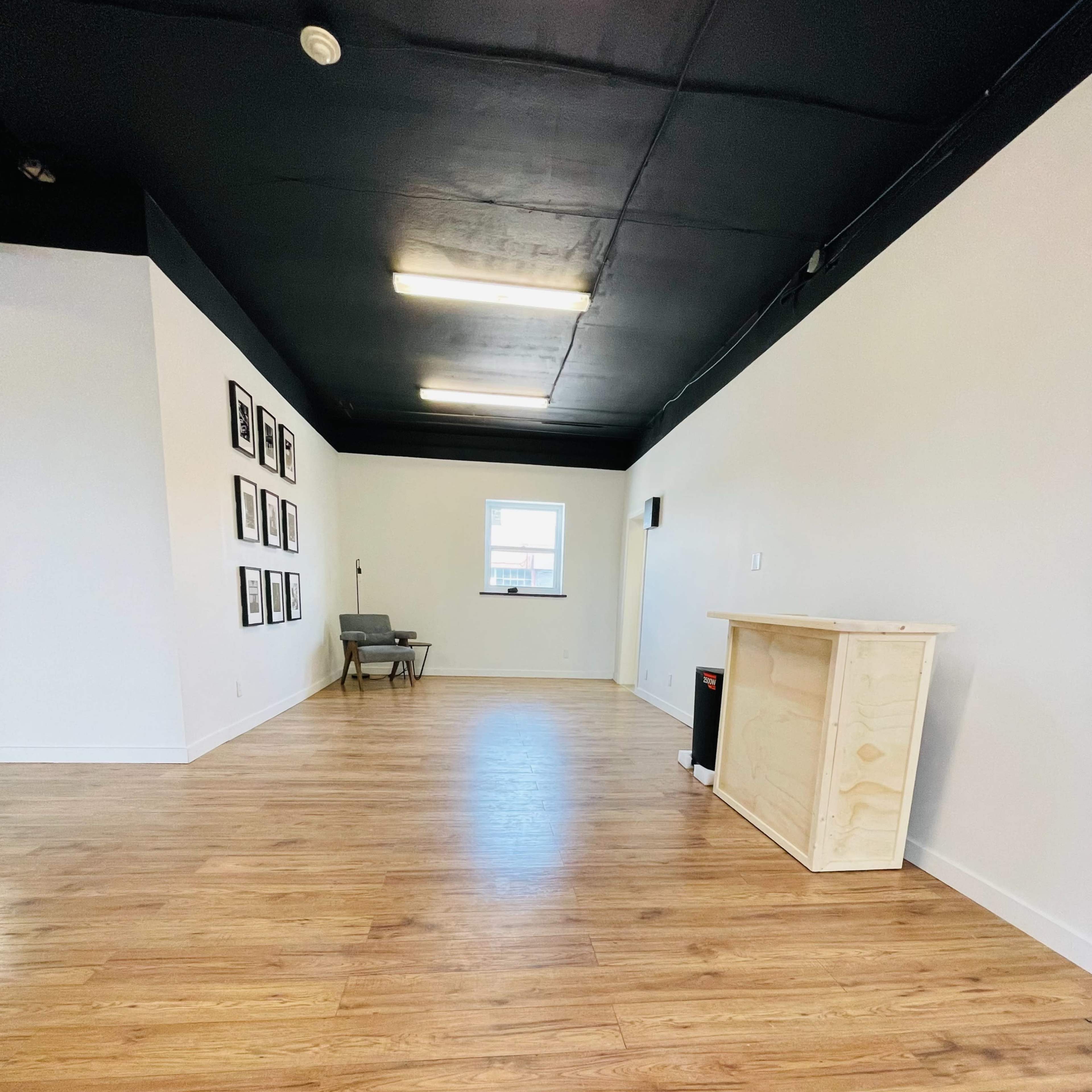 A spacious, empty room with wooden flooring, a wall of picture frames on one side, and a small wooden counter placed near the entrance.