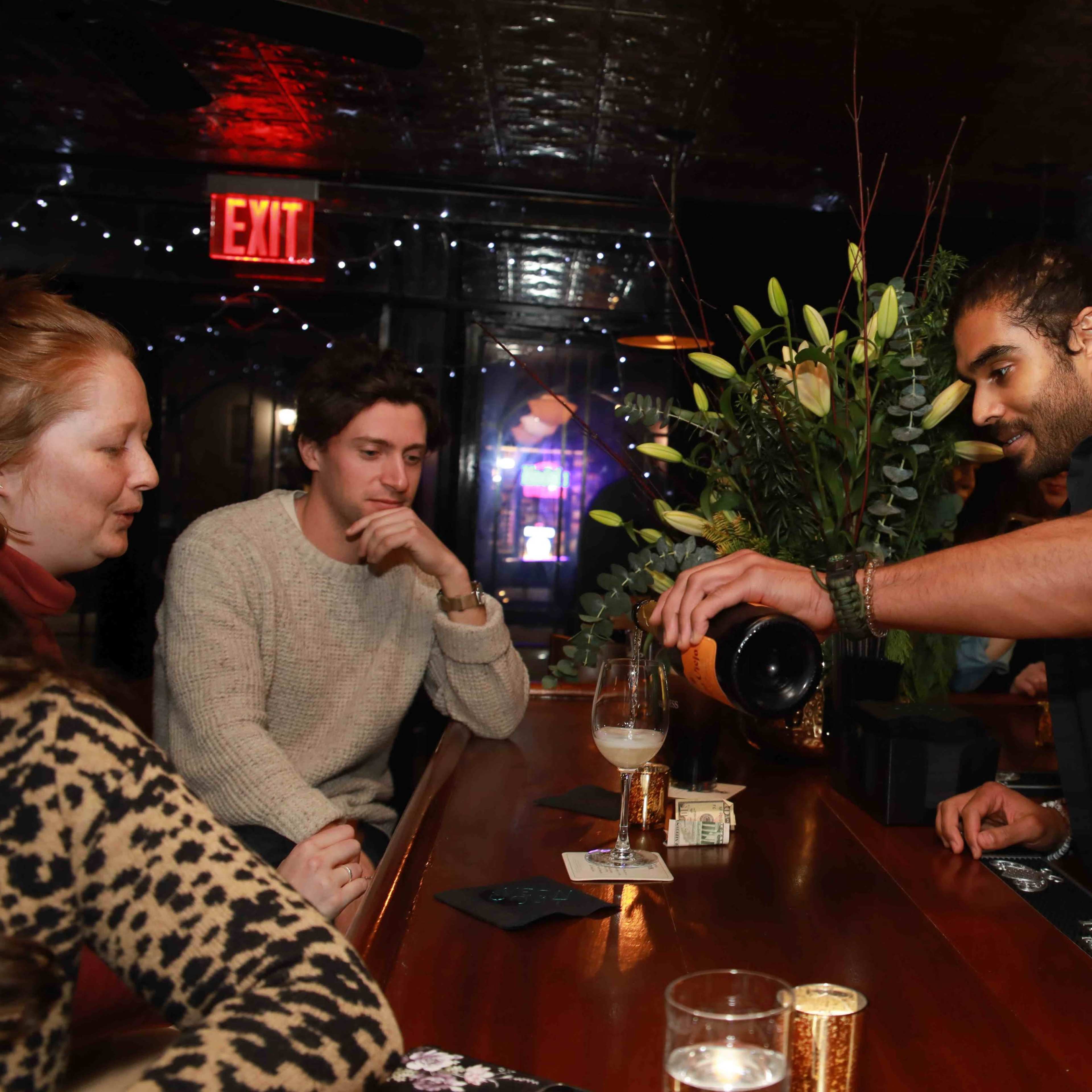 A bartender pours a drink for two seated customers at a warmly lit bar adorned with floral arrangements.