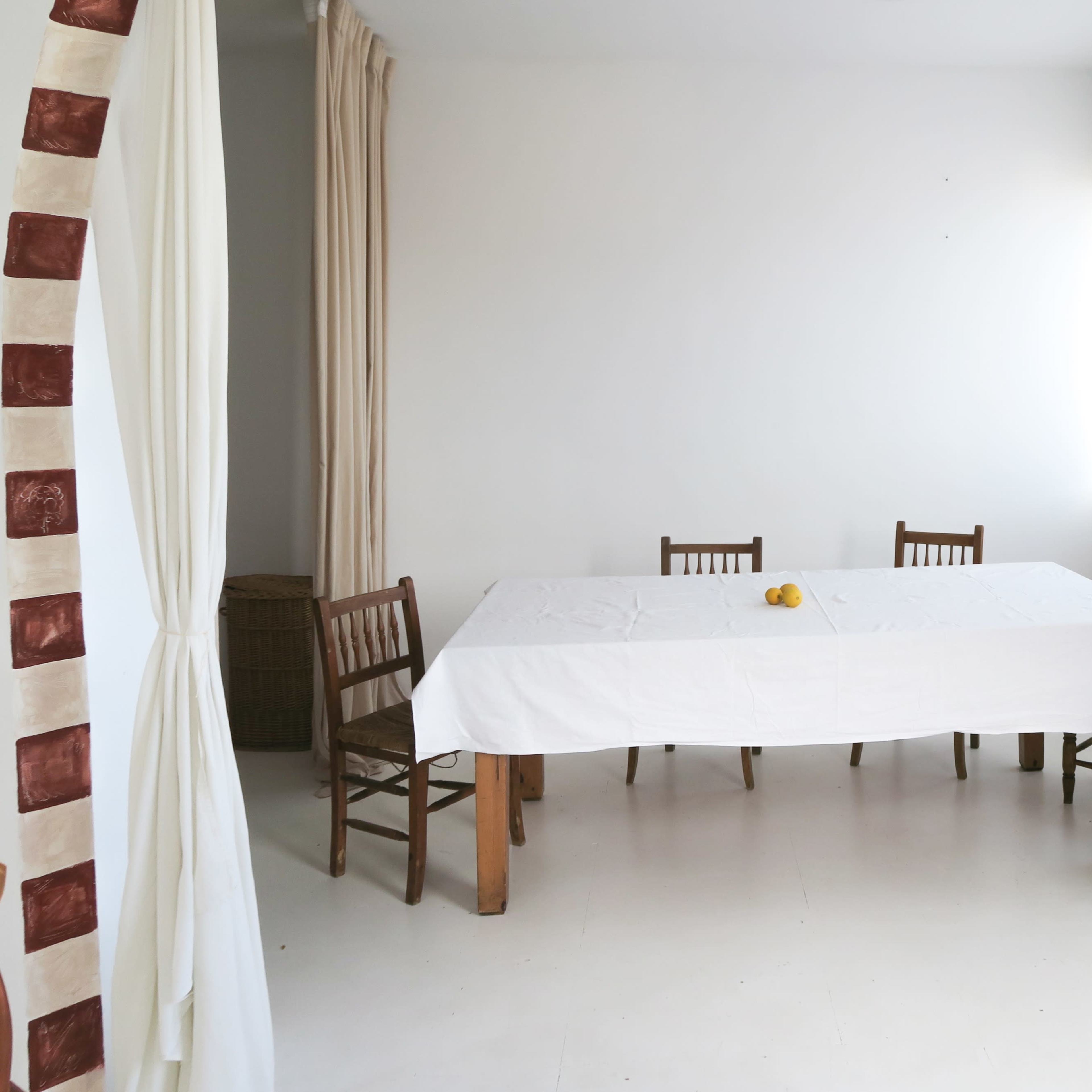A plain, bright room features a large white table surrounded by four wooden chairs, with a lamp and a small vase on a side table.