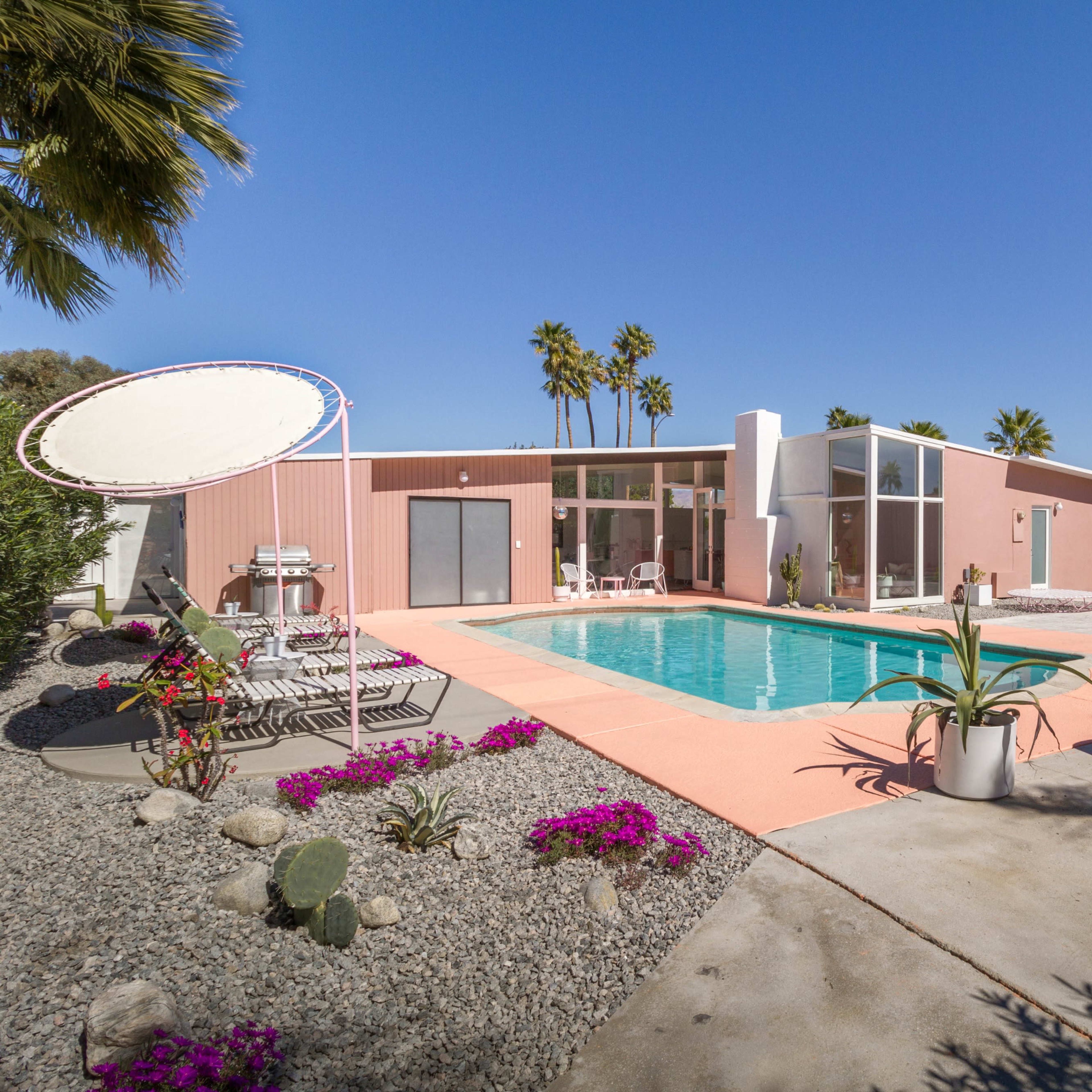 A modern pink and white house with a swimming pool surrounded by palm trees and landscaped gravel areas.