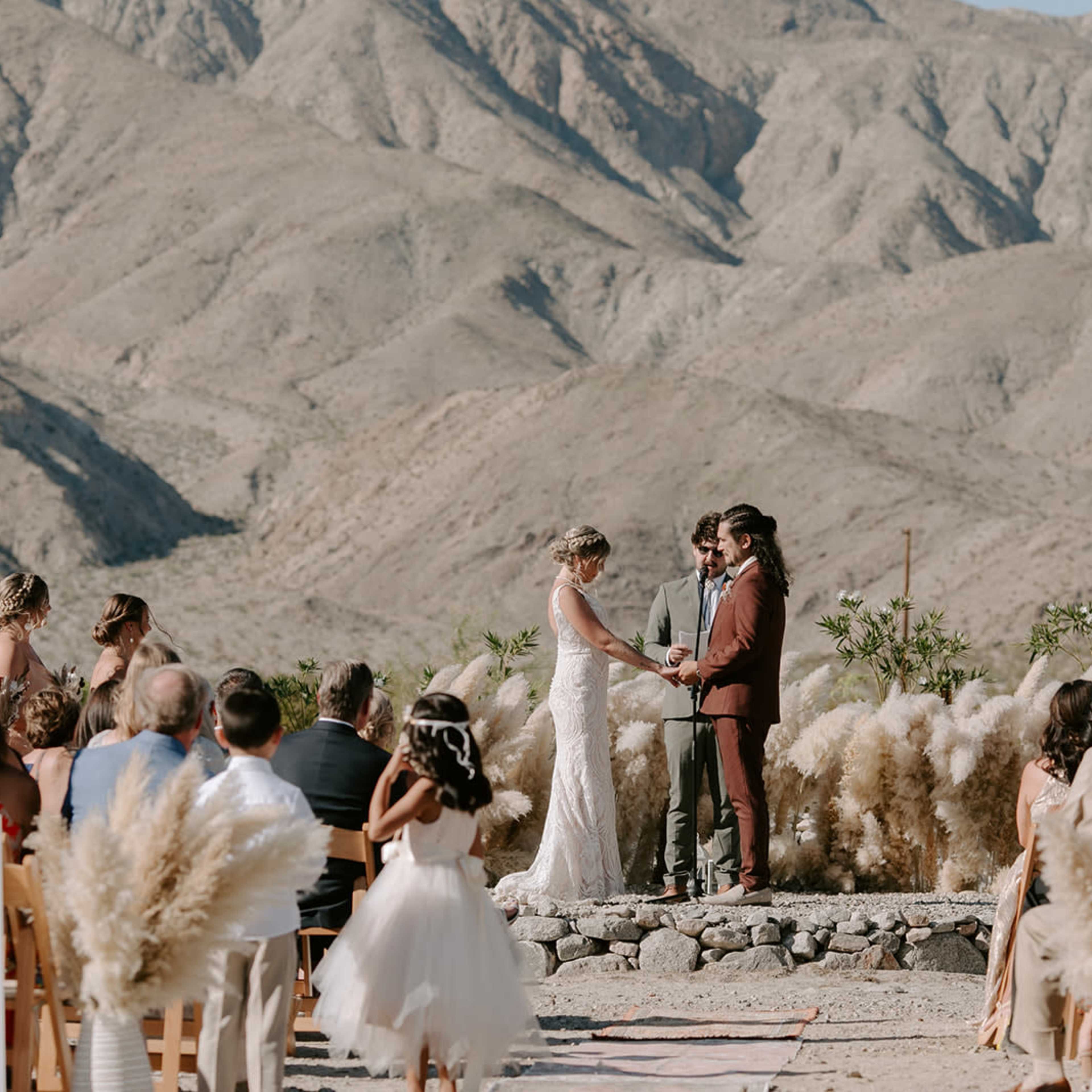 A couple stands on an altar surrounded by guests, with mountains in the background, during a wedding ceremony.