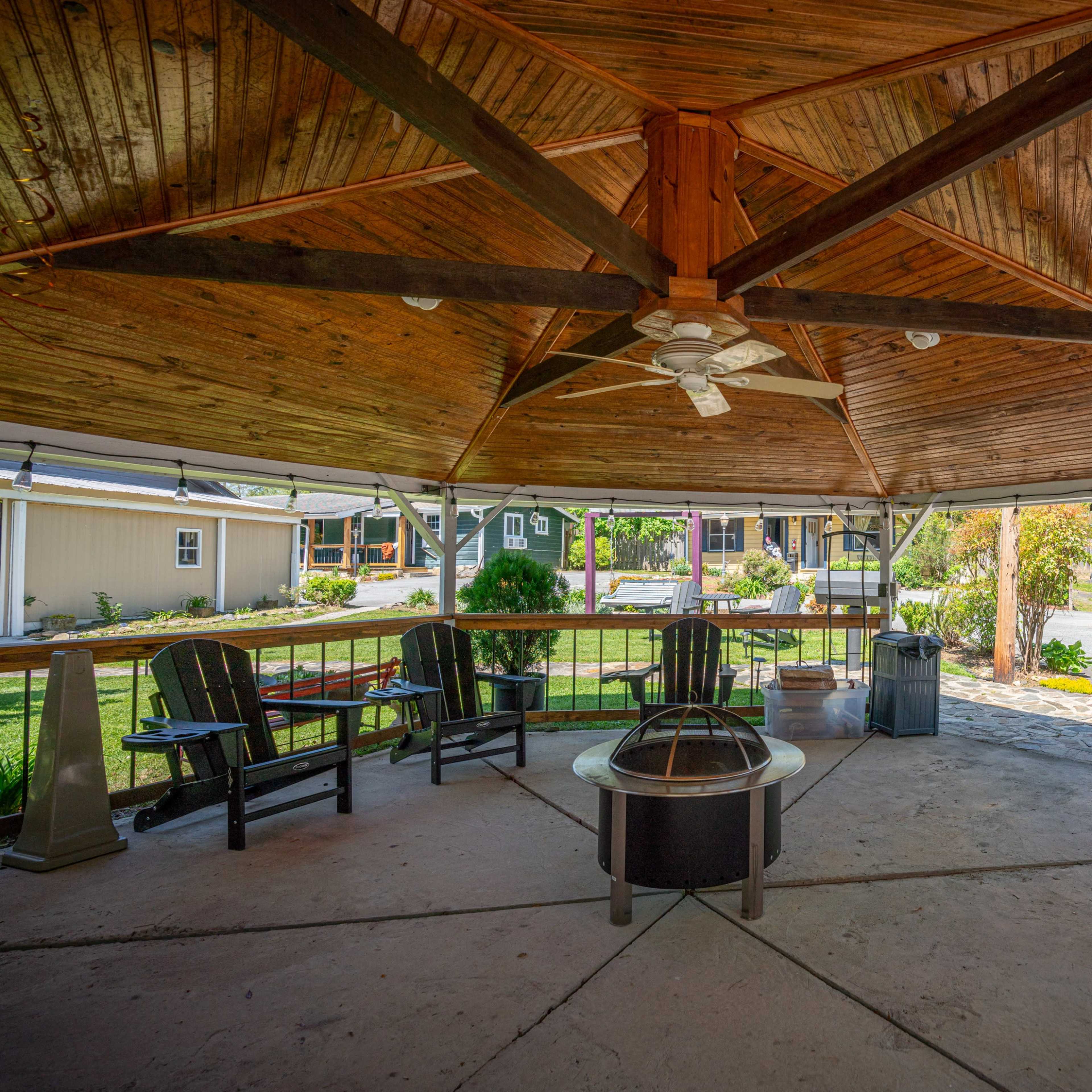 A wooden gazebo with a circular seating arrangement, a fire pit in the center, and a view of a landscaped area beyond.