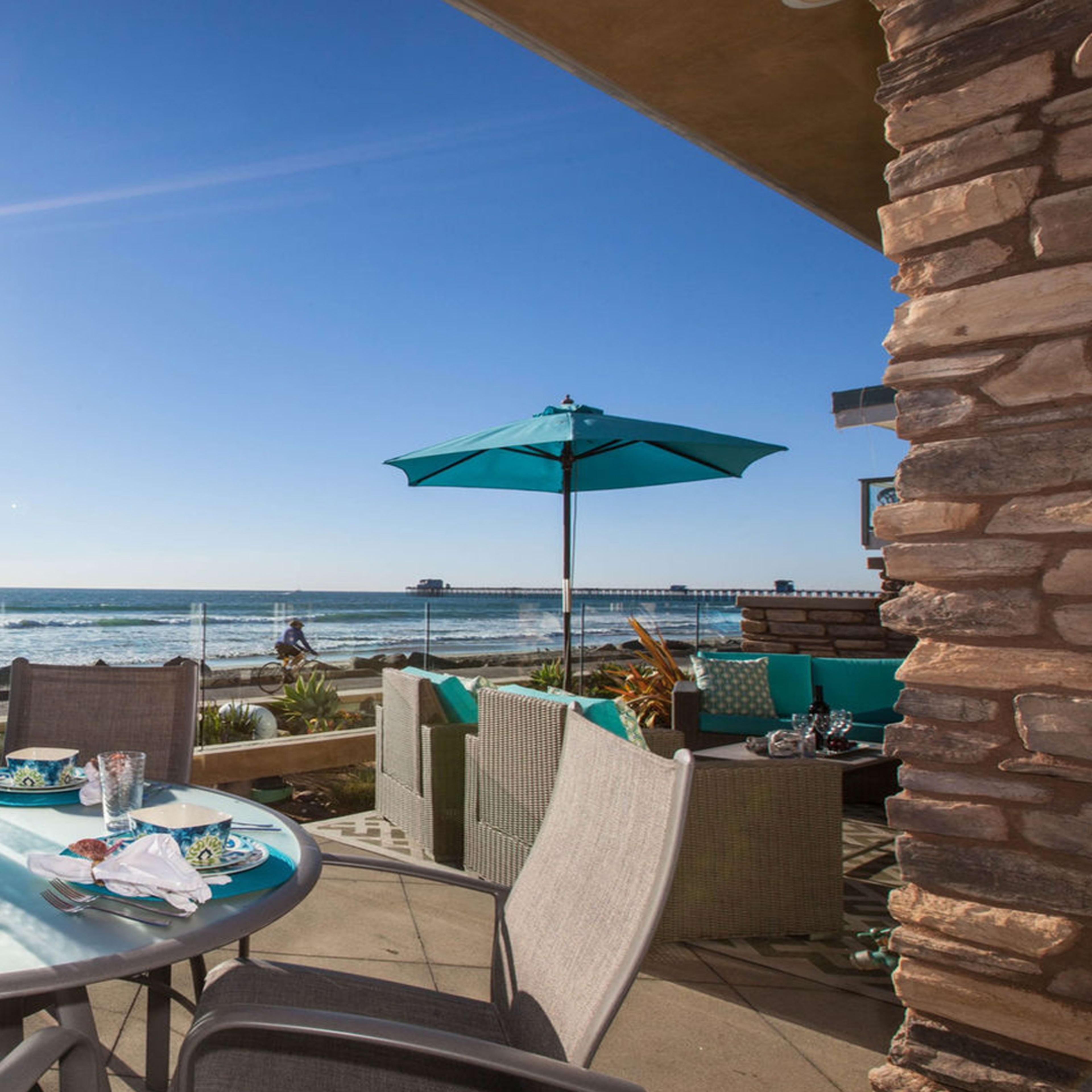 A dining table is set on a patio overlooking the ocean, with waves crashing in the background and a bright sun shining in the sky.
