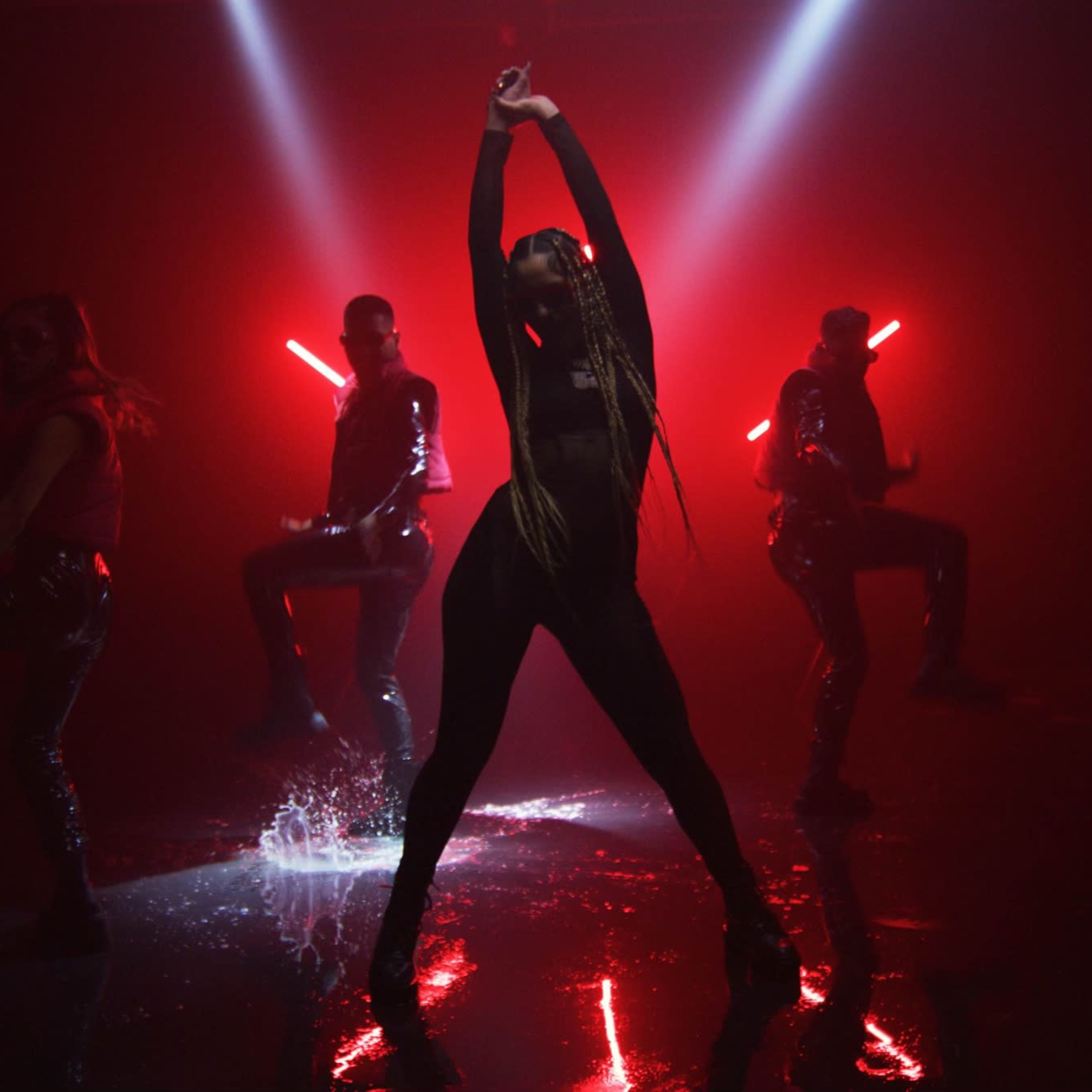 A dancer strikes a pose in the spotlight while three backup performers move in the background amidst dramatic lighting and reflections on a wet surface.