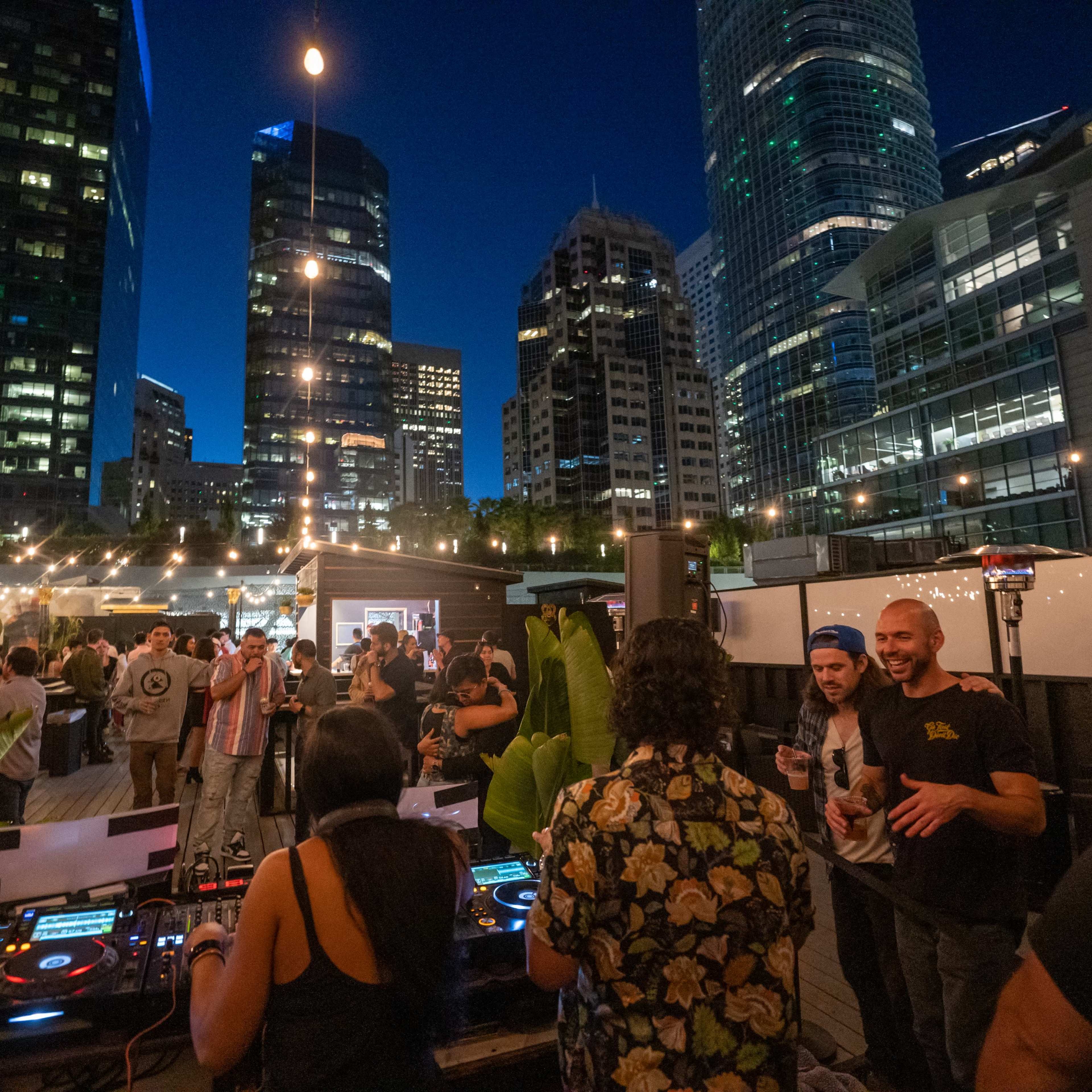 A rooftop party is taking place at night with people dancing and socializing, while a DJ plays music in front of tall city buildings.