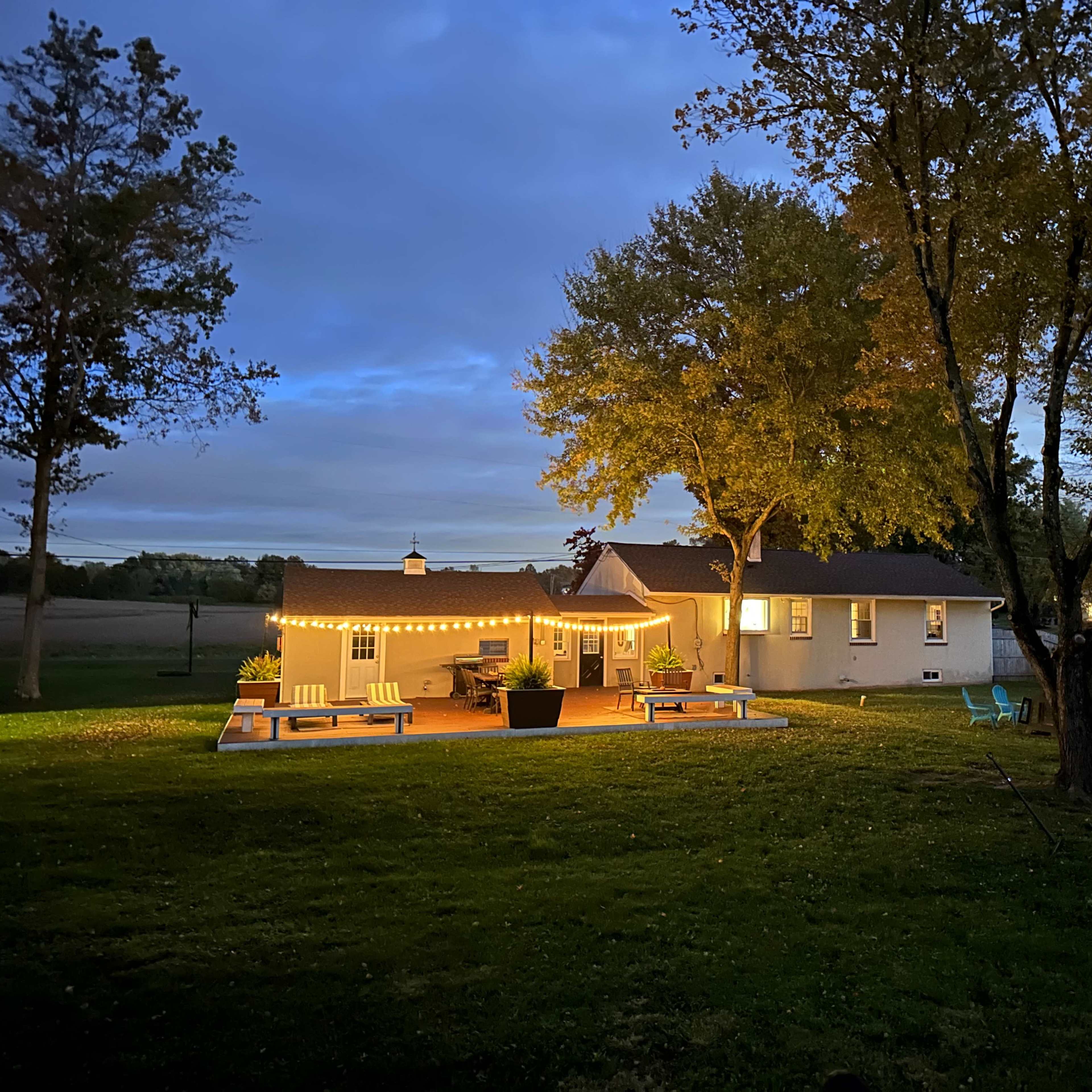 A well-lit house with string lights sits in a grassy yard surrounded by trees as dusk settles.