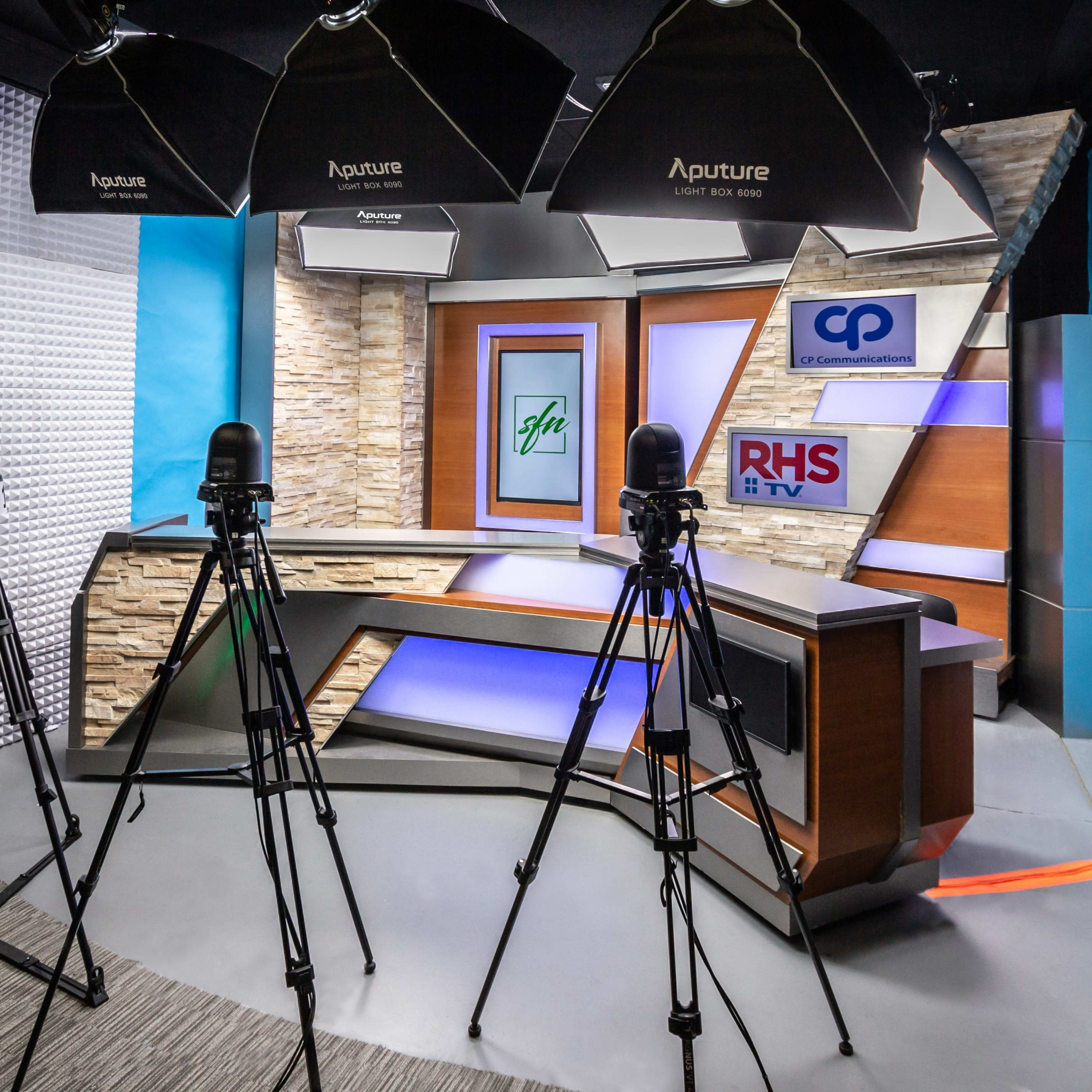 A television studio setup with three tripods positioned in front of an anchor desk, featuring a branded backdrop and studio lighting.