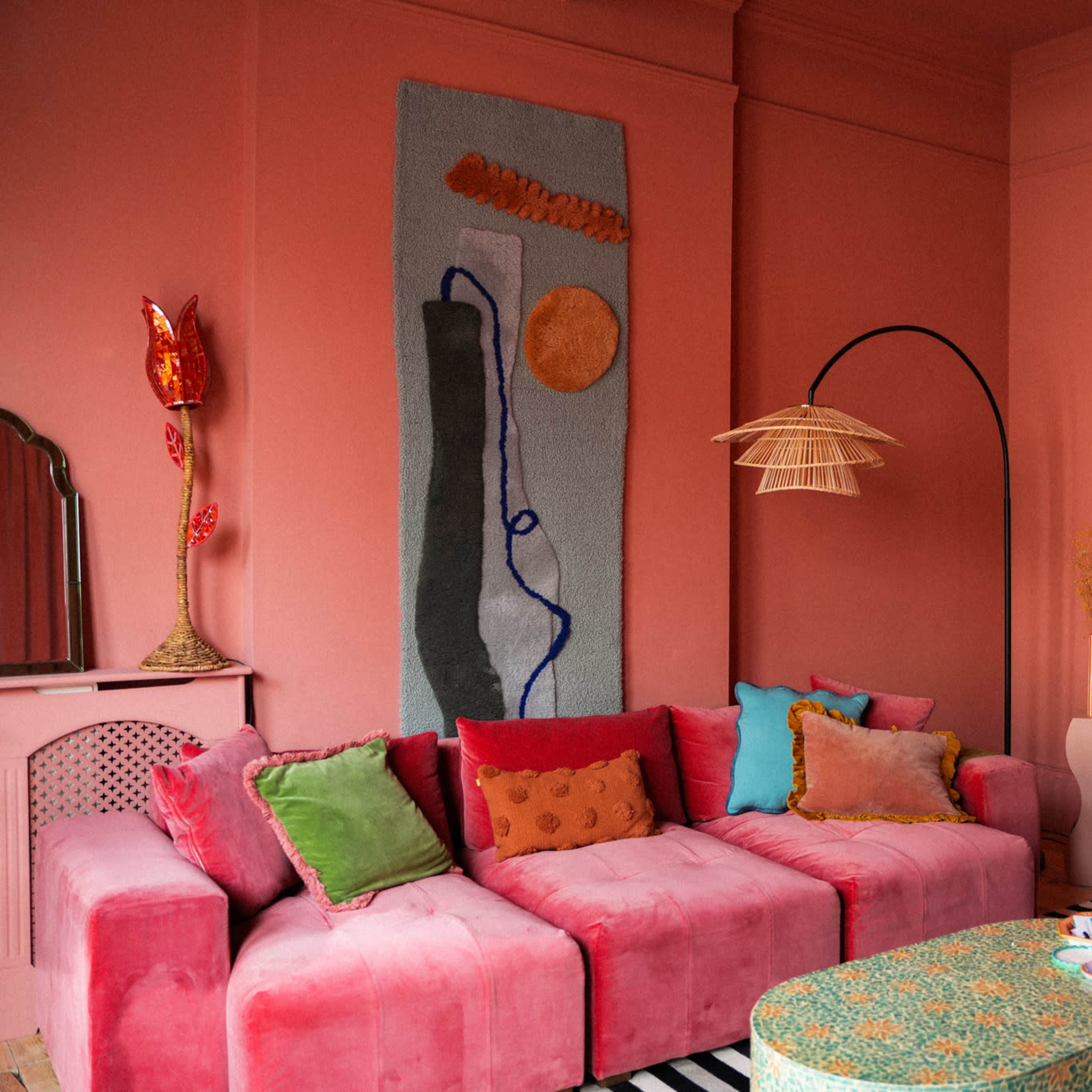 A colorful living room with a pink wall, a plush sectional sofa adorned with various cushions, a wooden shelf displaying decorative items, and a mural hanging on the wall.