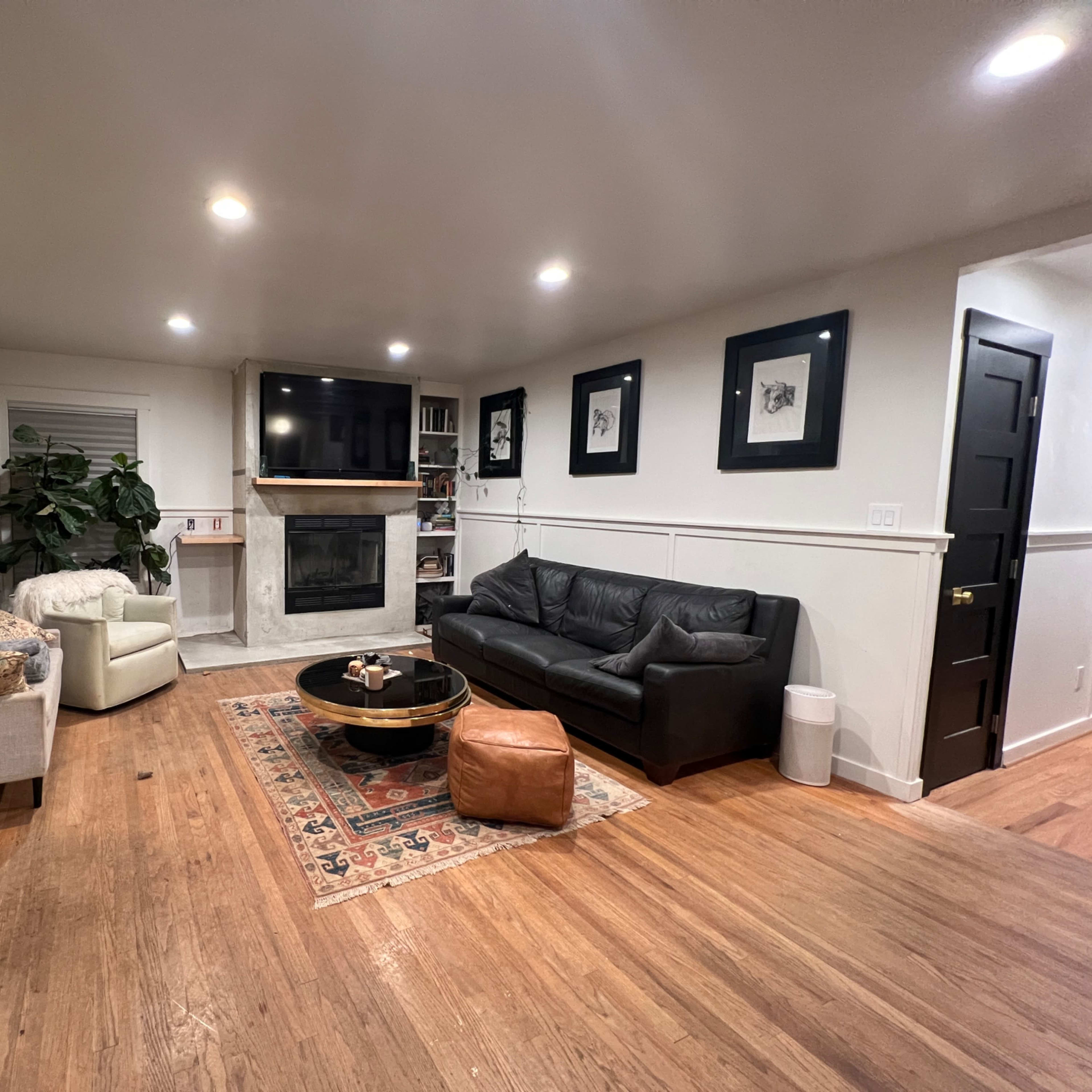 The living room features a leather sofa, a round coffee table, a flat-screen TV above a fireplace, and light wood flooring with a decorative rug.