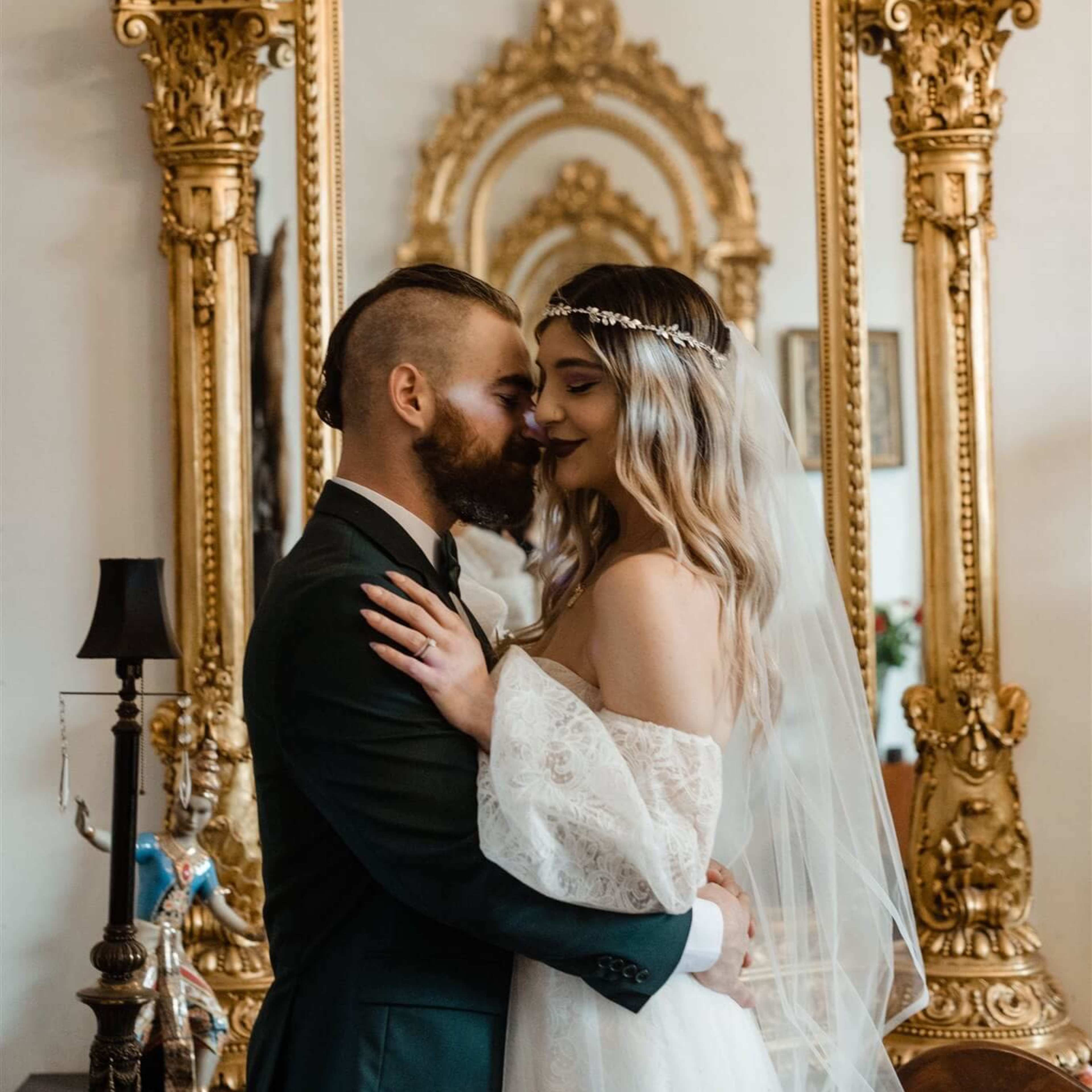 A couple embraces in front of an ornate, gilded mirror in a decorated interior.