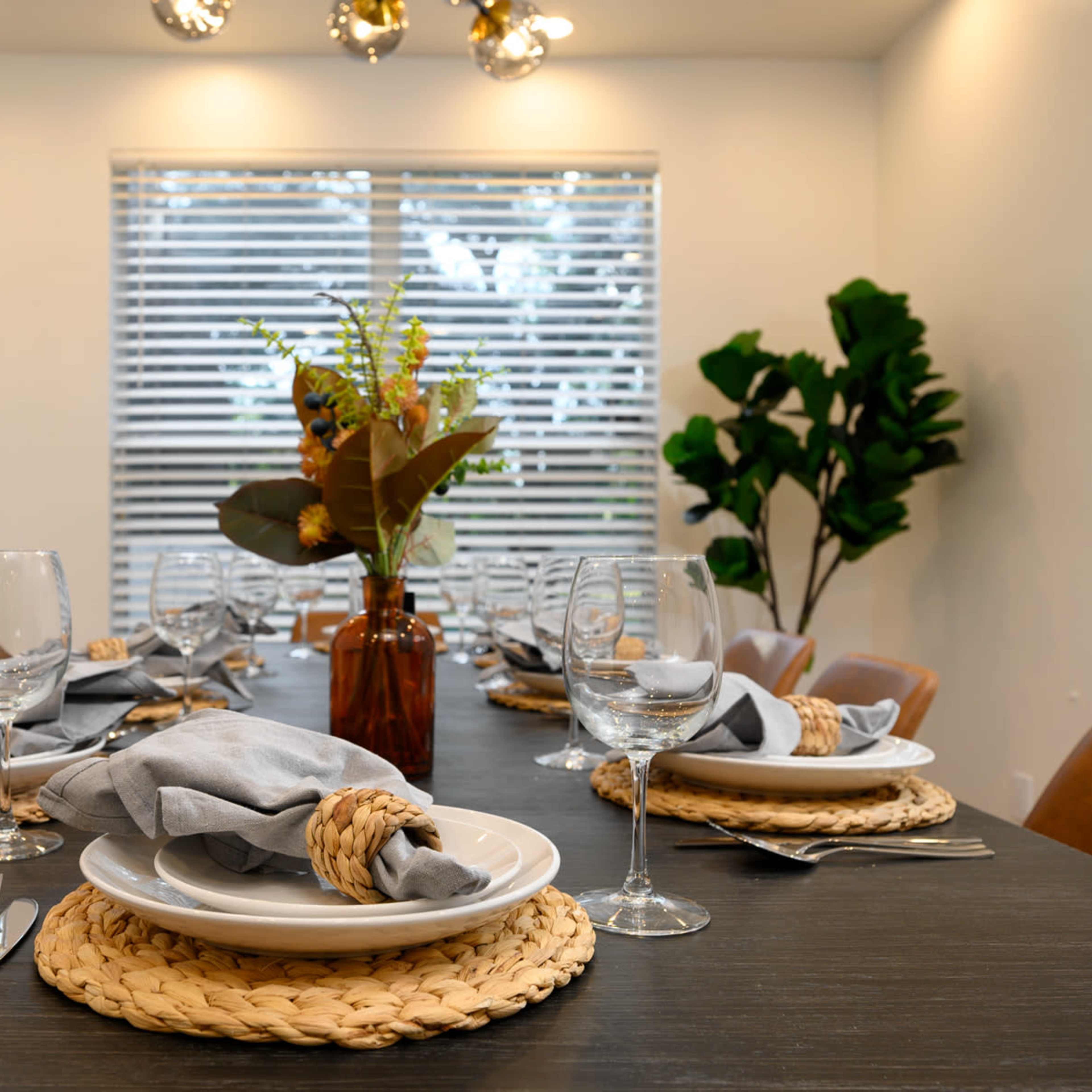 A dining table is set with glassware and neatly arranged plates, surrounded by chairs, and features a vase with flowers in a well-lit room.