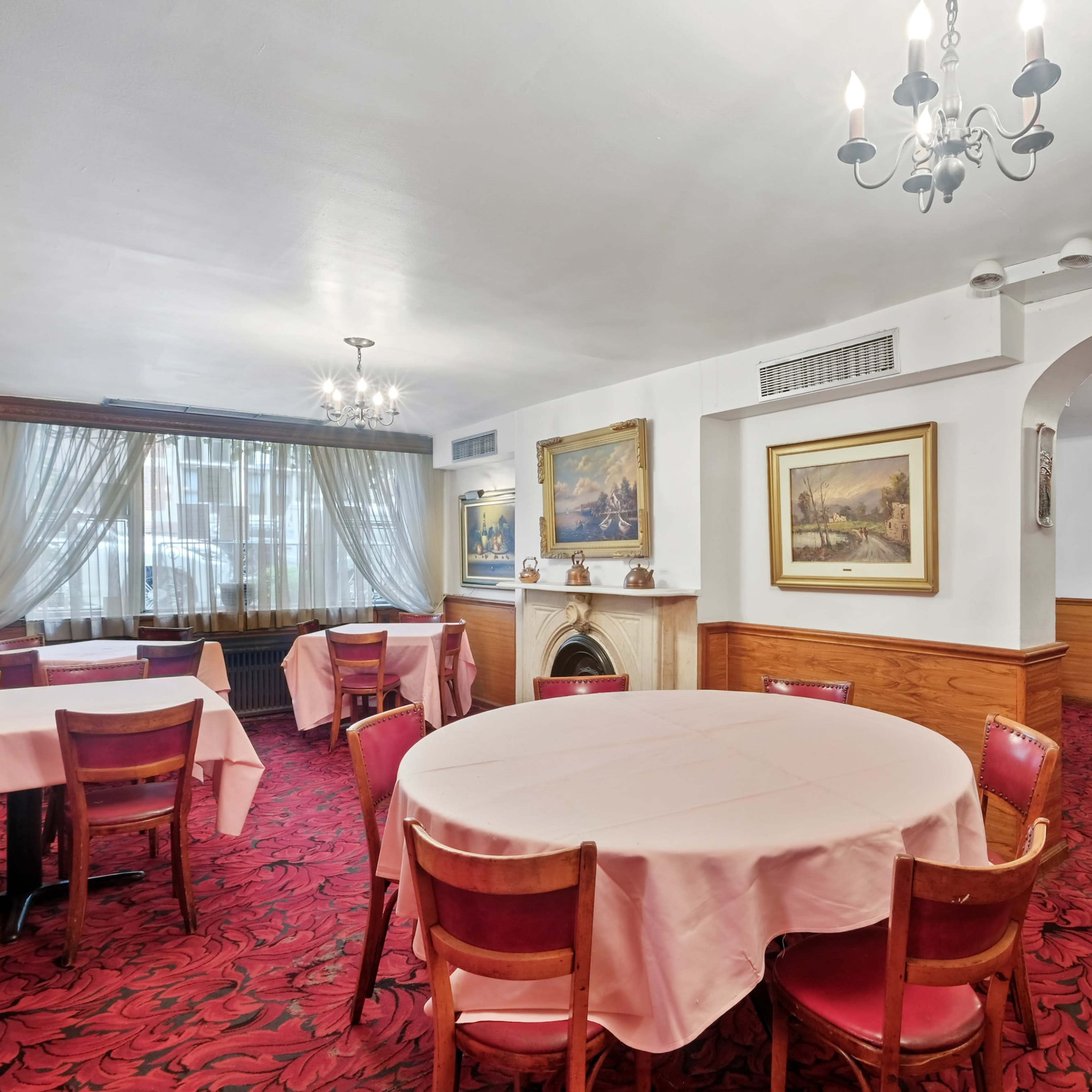 An elegantly furnished dining room with round tables covered in pink tablecloths, red patterned carpeting, and framed paintings on the walls.