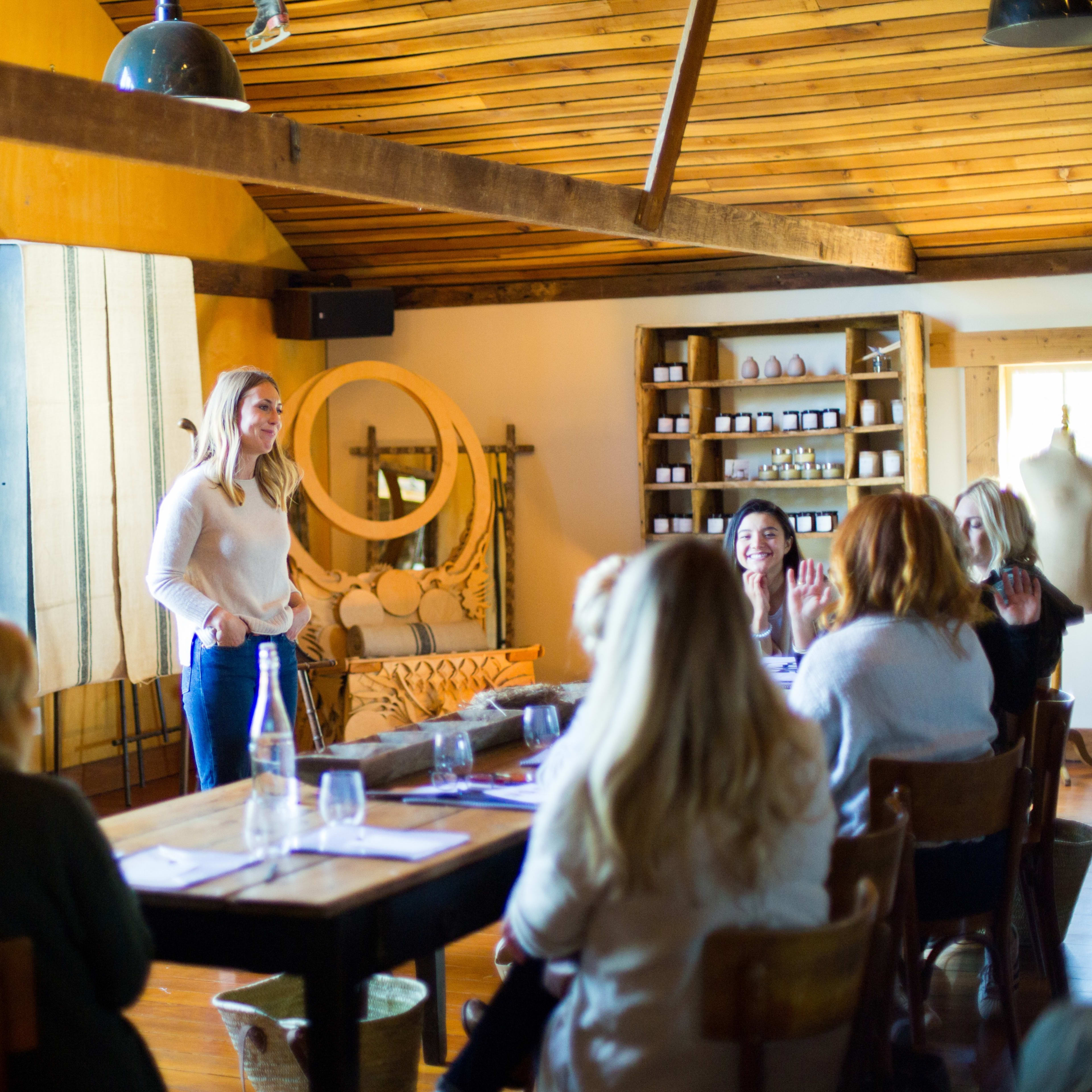 A woman stands in front of a group of seated individuals in a bright, rustic meeting space with wooden beams and shelves.