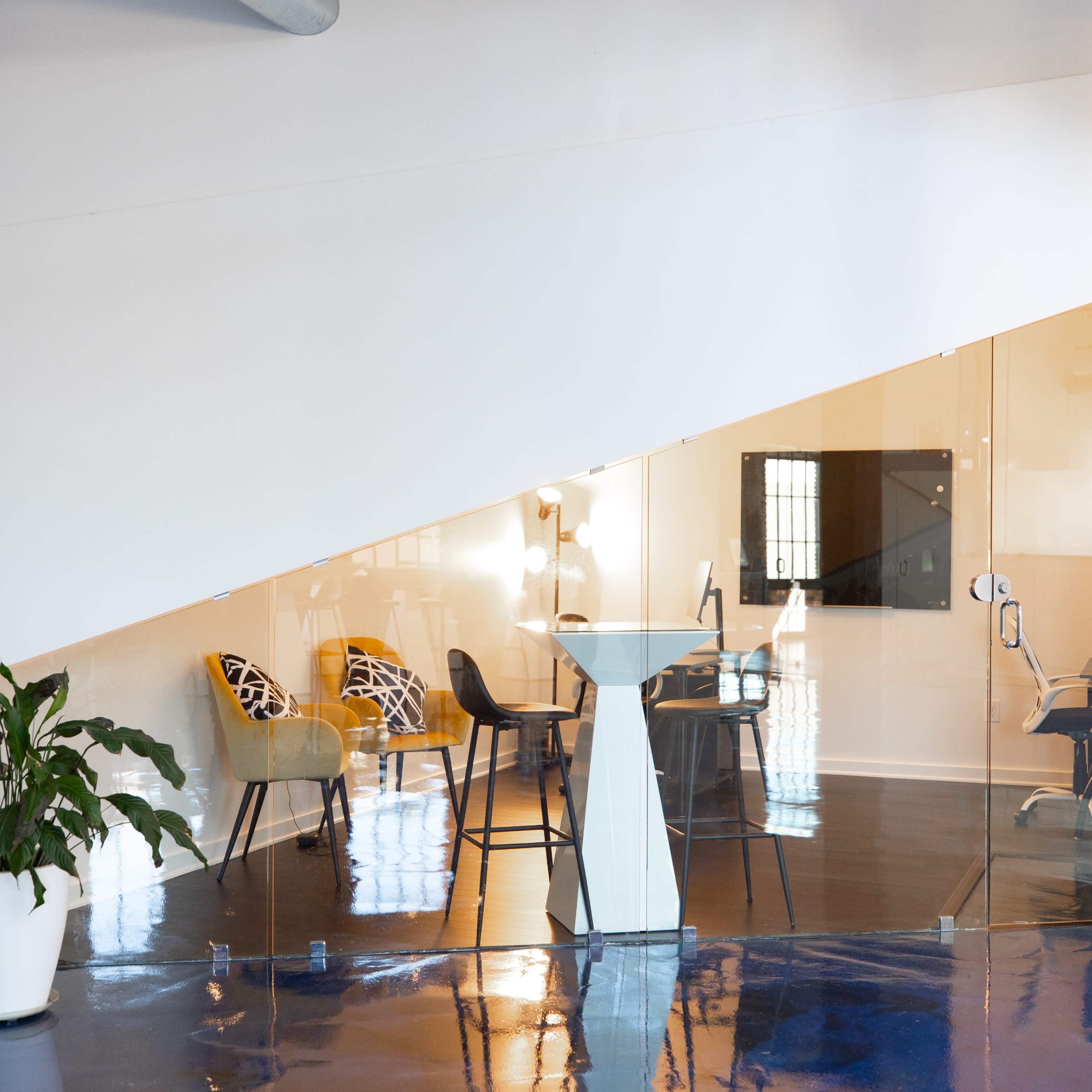 A modern office space features a glass-walled meeting area beneath a staircase, with seating arrangements and a potted plant in the foreground.