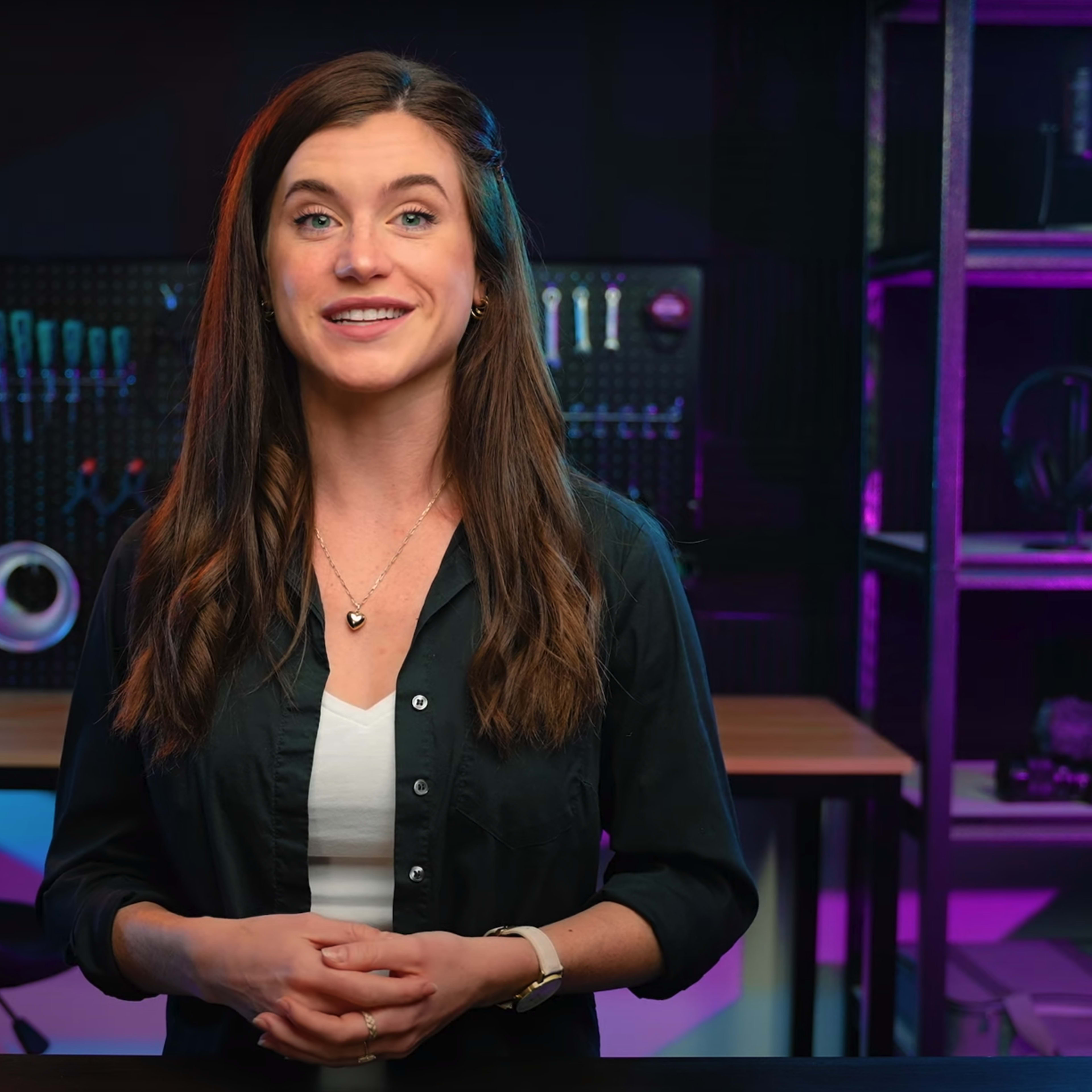 A woman stands in front of a shelf filled with various audio and video equipment, smiling at the camera.