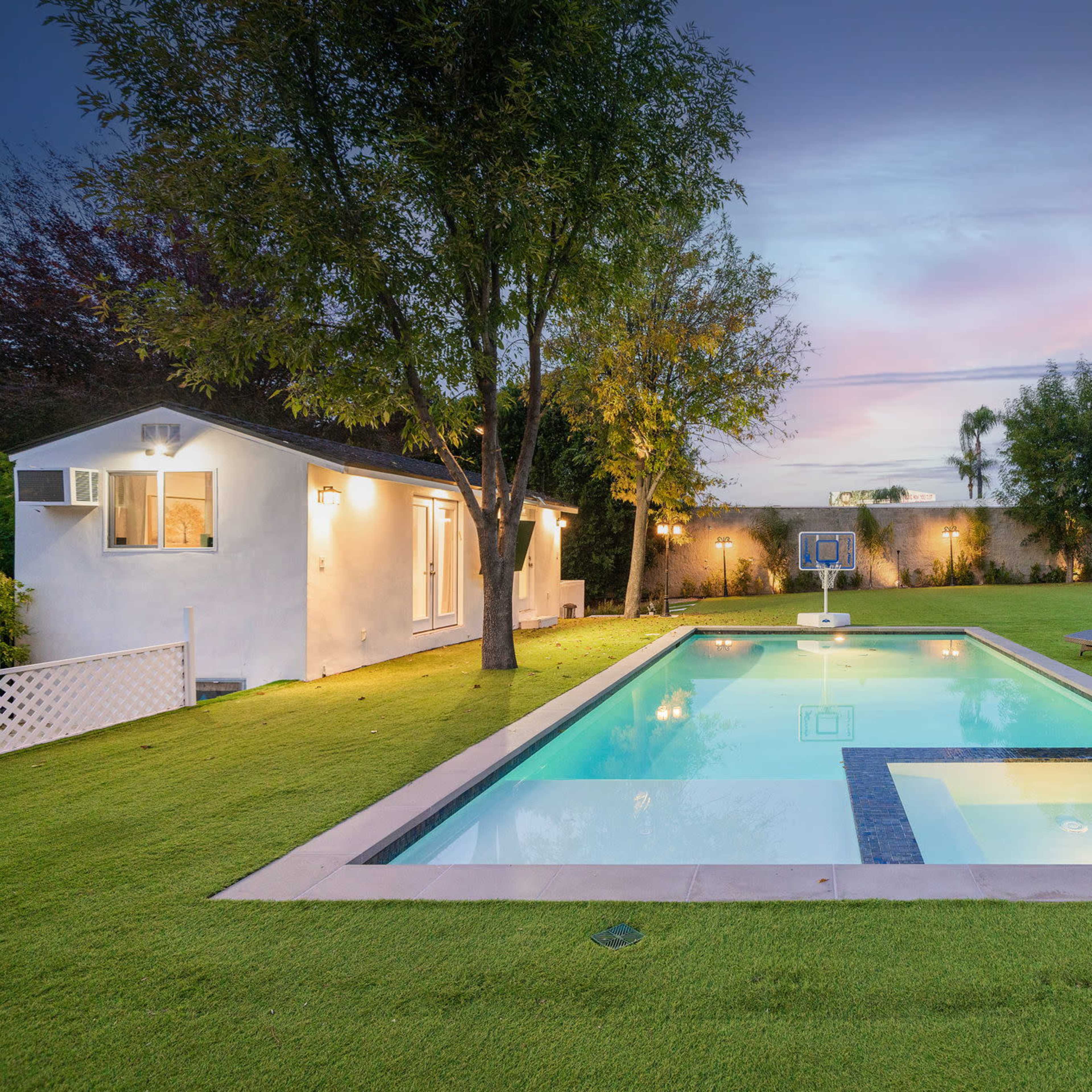 A rectangular swimming pool surrounded by a lush green lawn, with two lounge chairs nearby and a small building in the background.