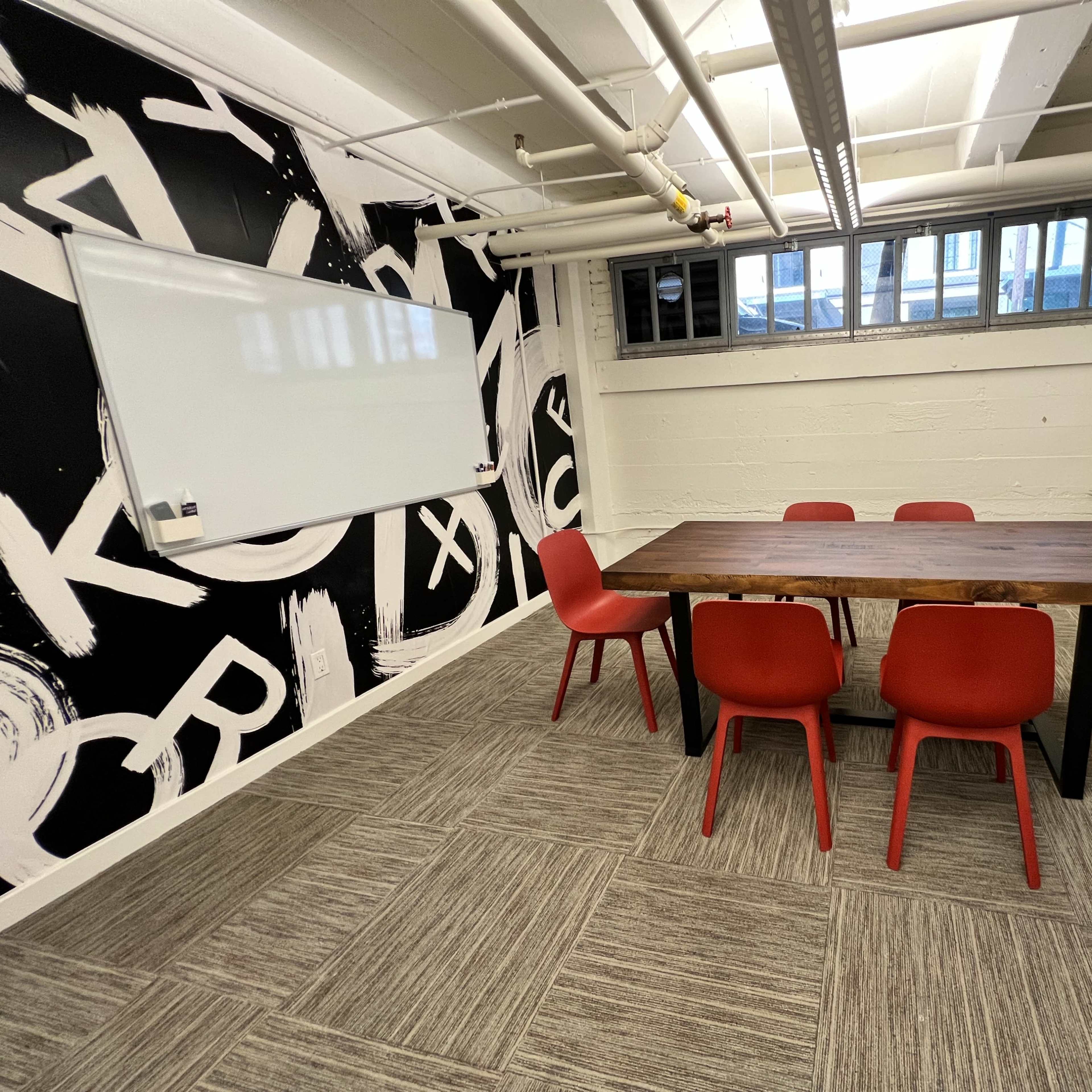 A conference room features a wooden table surrounded by six red chairs, with a whiteboard on one wall and a graphic black-and-white mural on another.