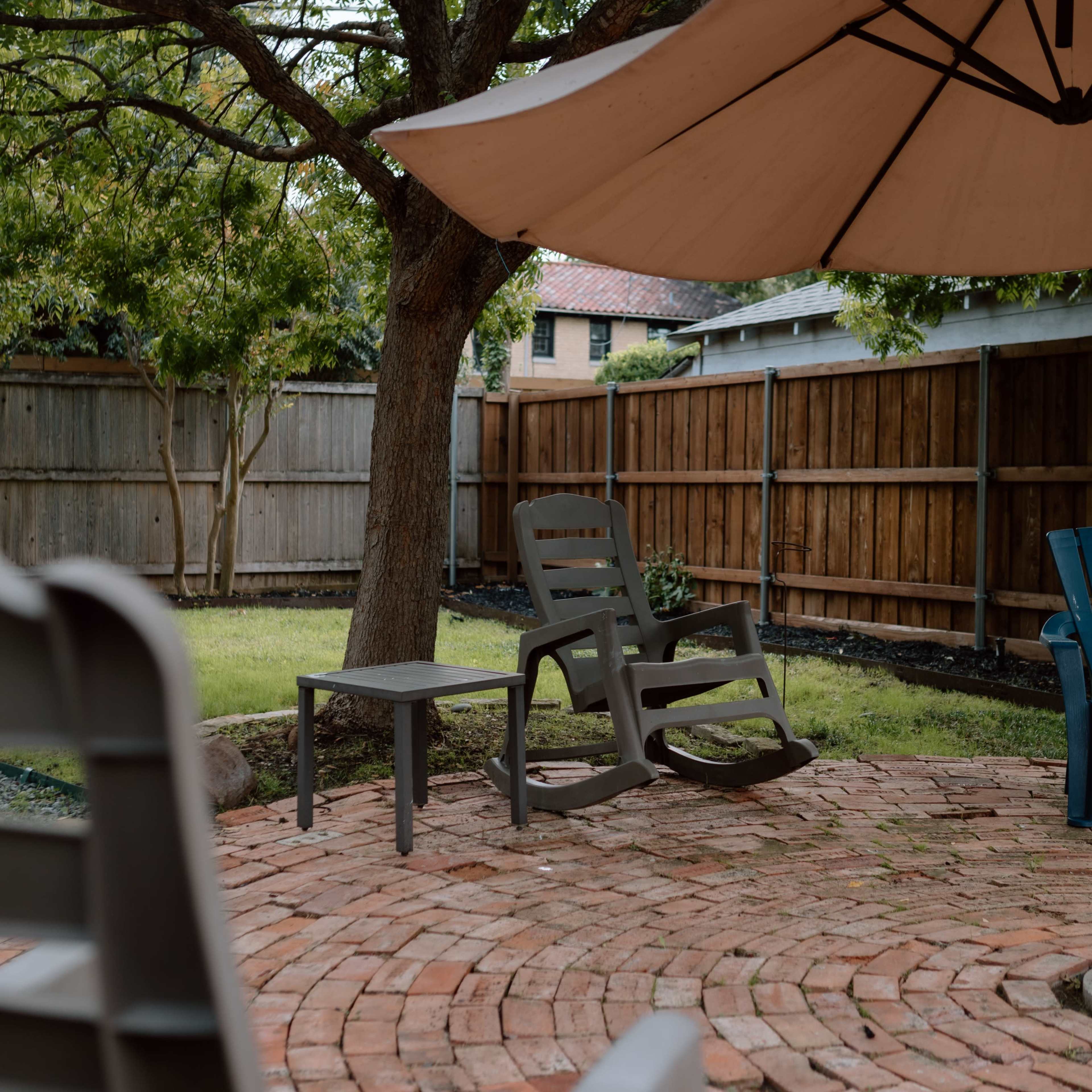 A backyard patio with brick flooring, a large umbrella, and a few patio chairs surrounded by trees and a wooden fence.