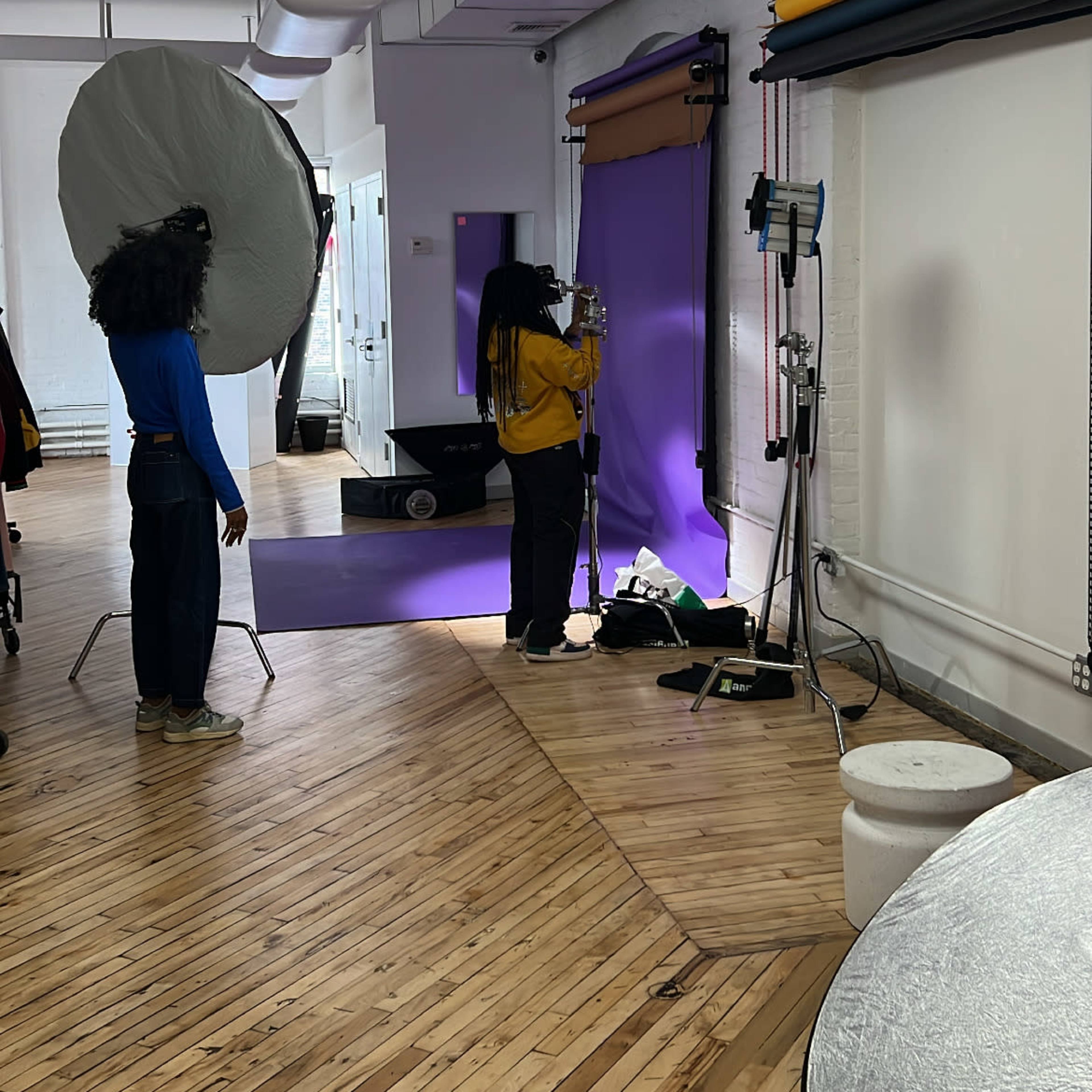 A photography studio with a person adjusting equipment in front of a purple backdrop, while another individual stands nearby near large reflectors.