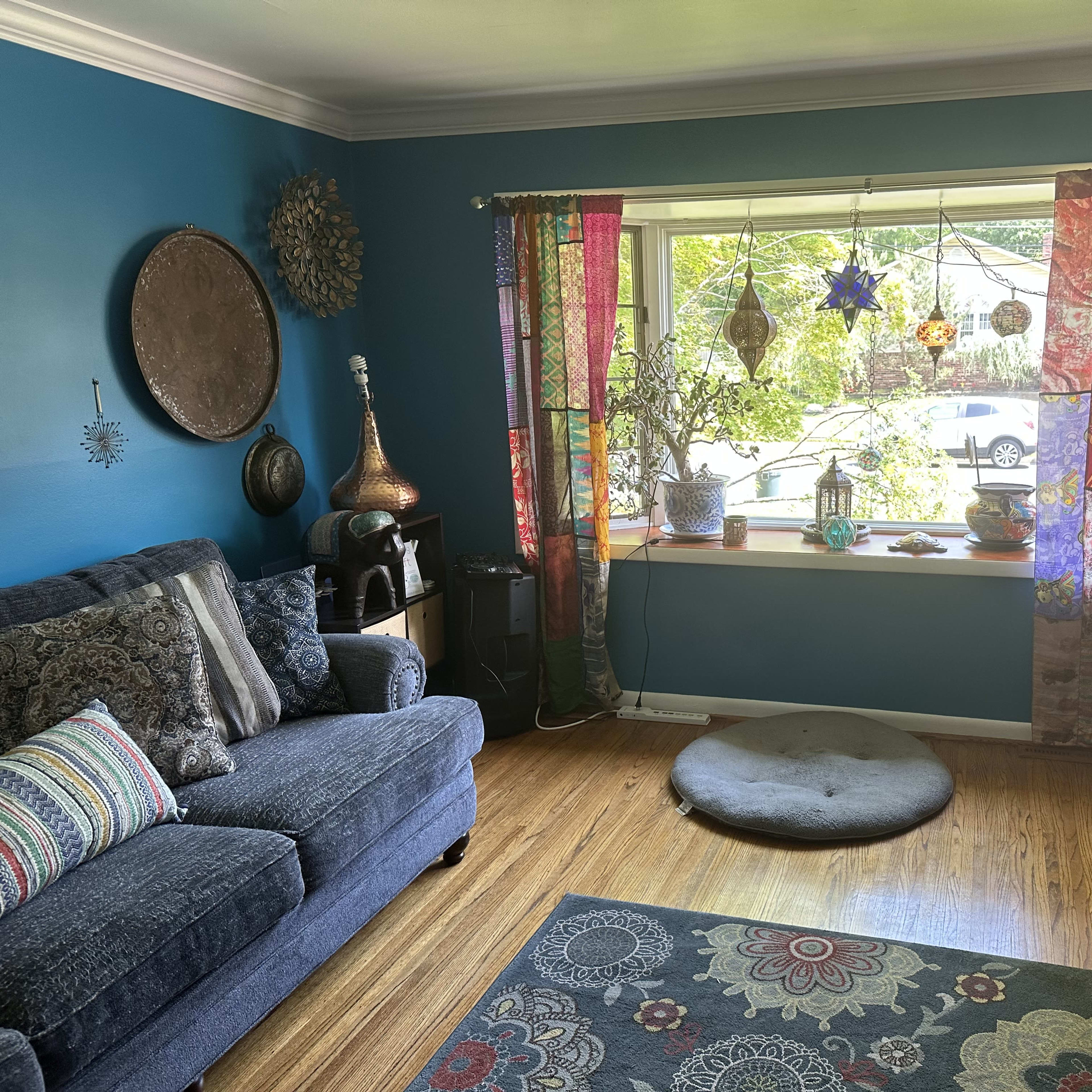 A living room with a blue wall, a sofa, colorful curtains, and a window displaying plants and a view of the outdoors.