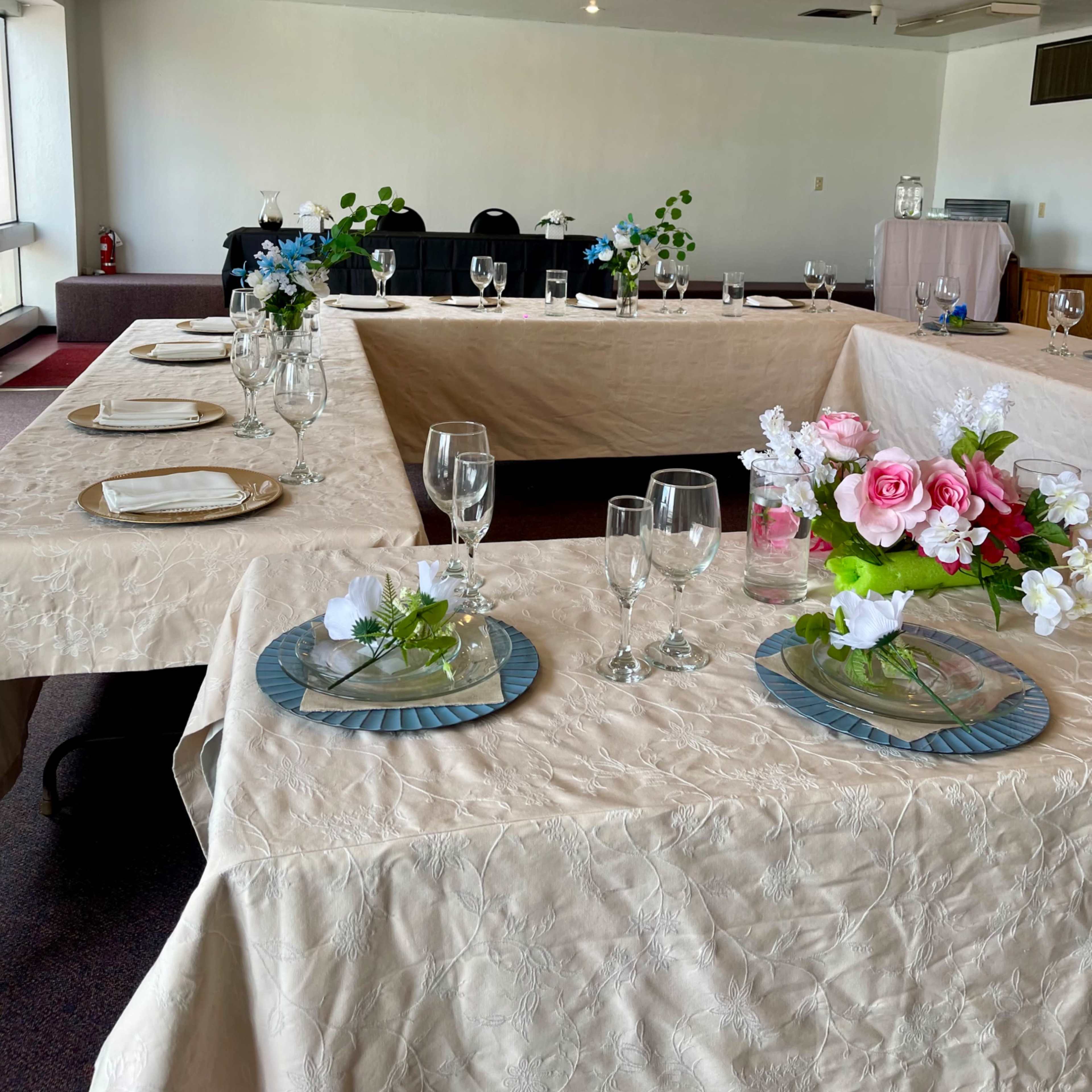 A dining area set with tables covered in light-colored tablecloths, each adorned with centerpieces of flowers and glassware.