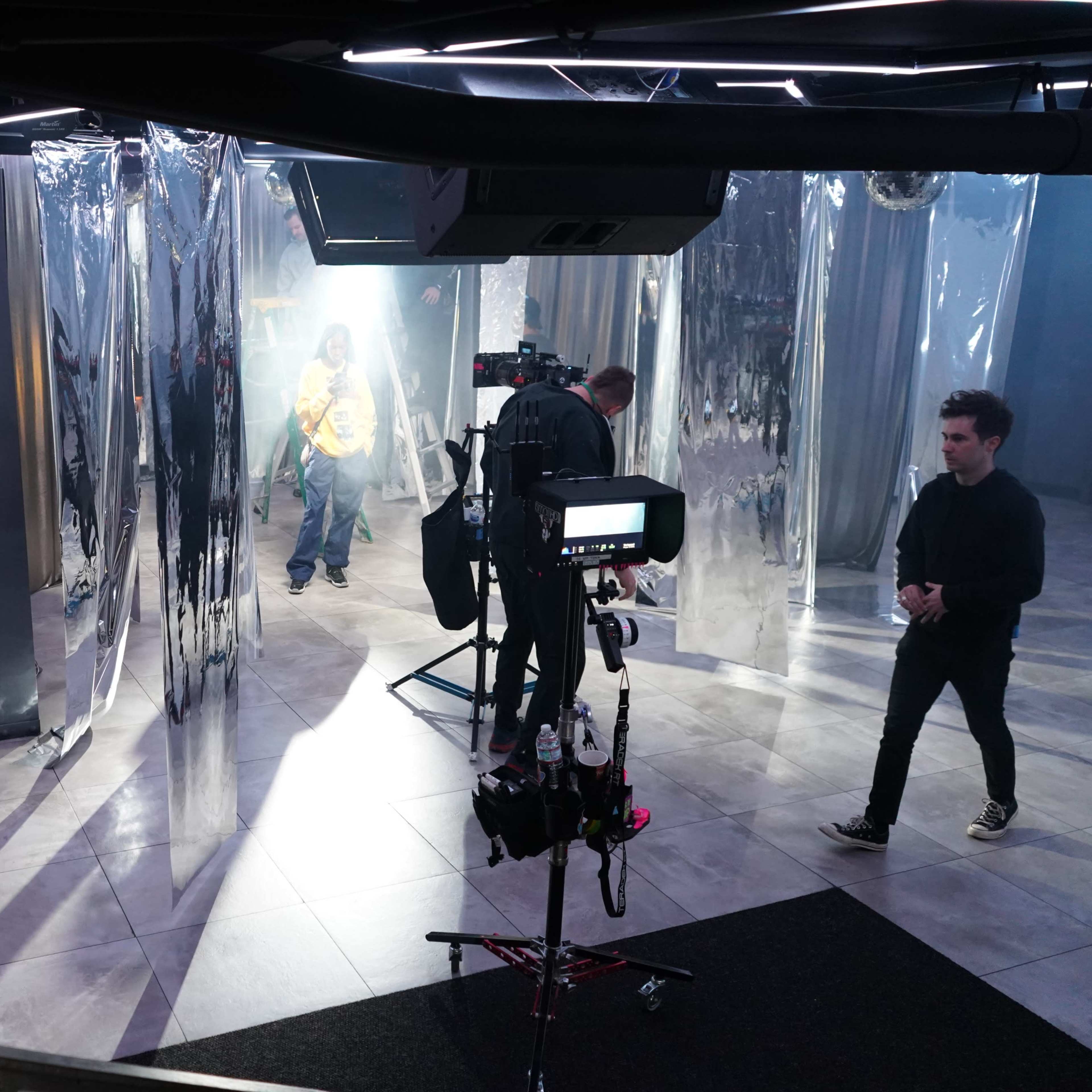 The scene shows a film set with crew members working around transparent plastic drapes and cameras positioned for a shoot.