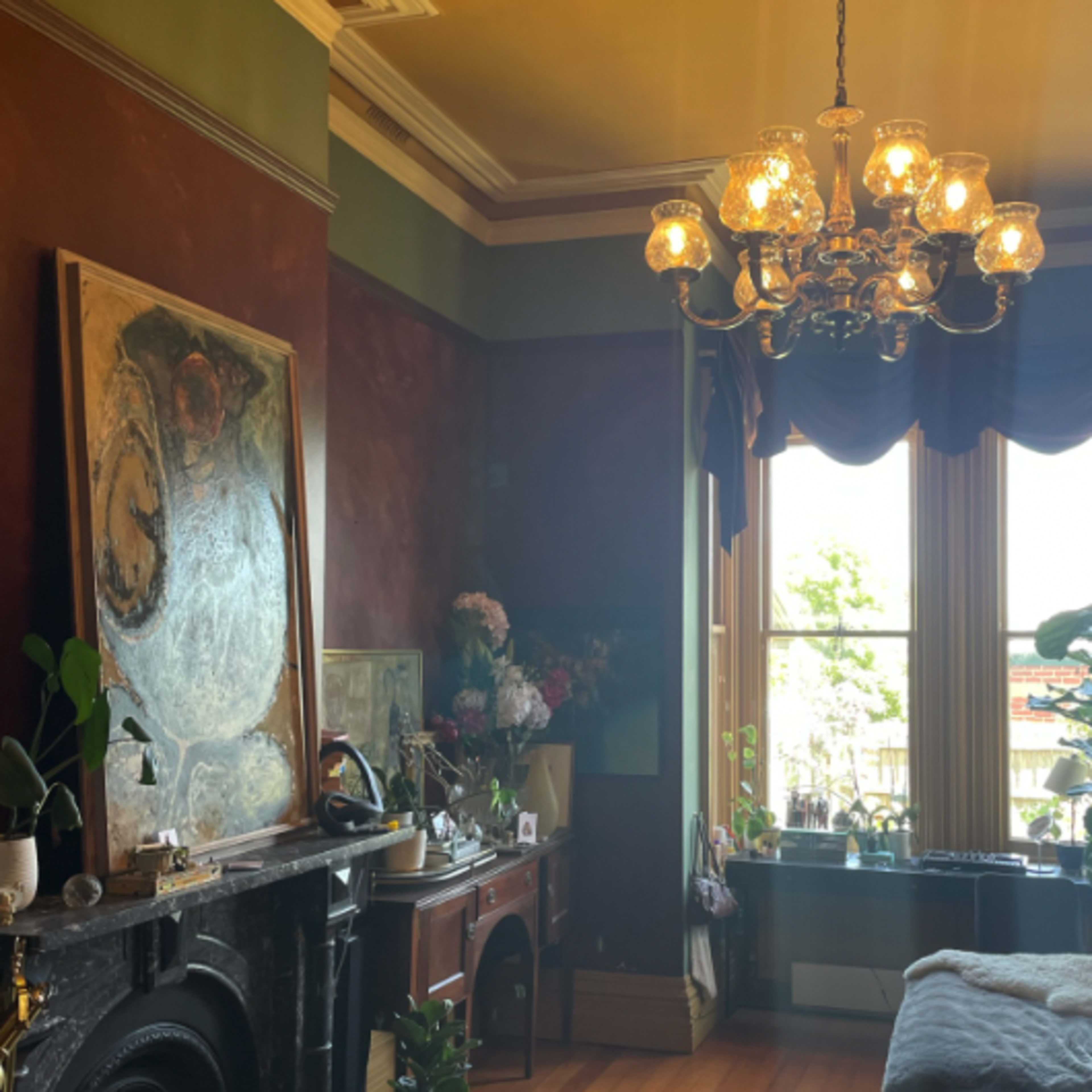 A well-lit room featuring a chandelier, large windows with drapes, a fireplace, and various plants and decorative items.