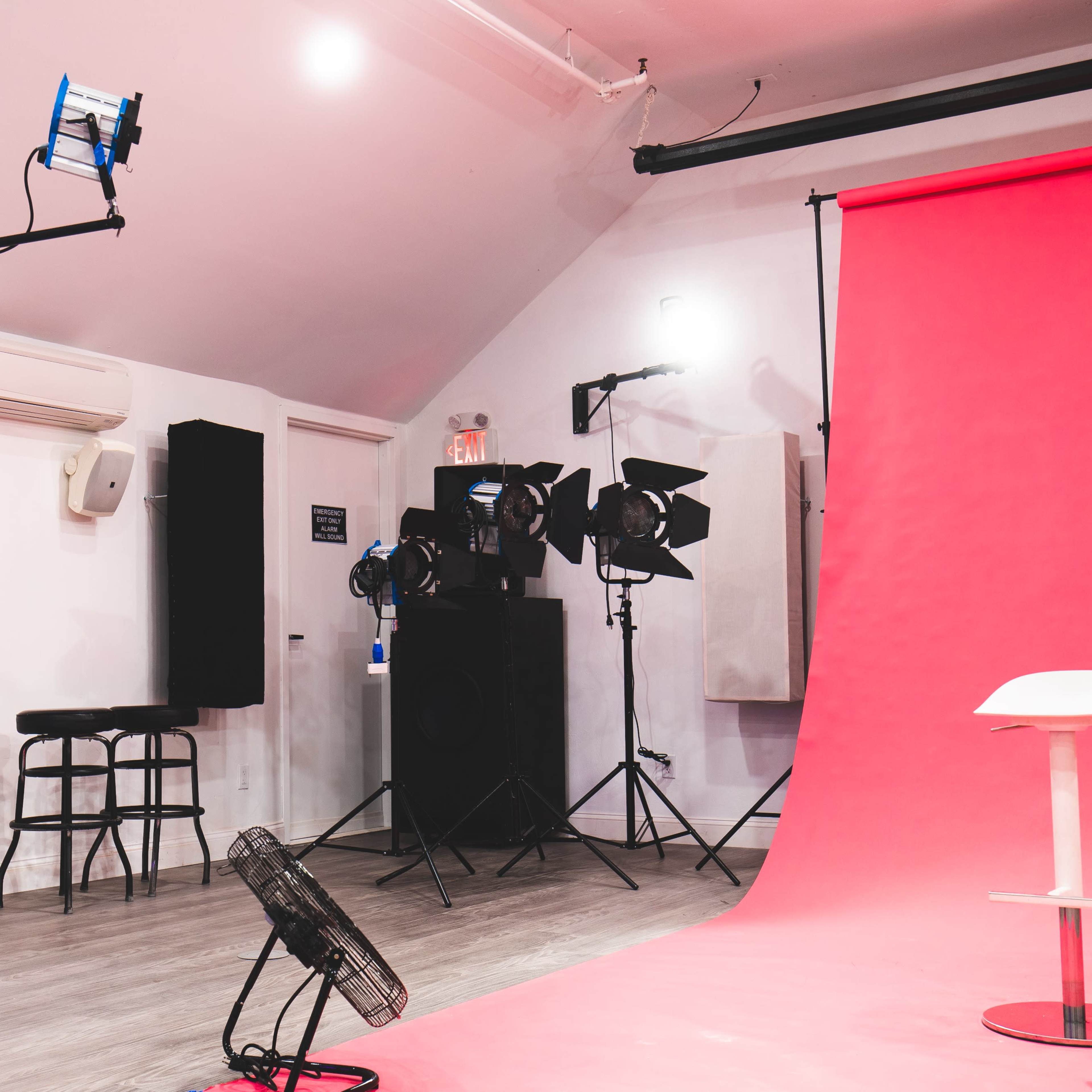 This image features a photo studio with a pink backdrop, studio lights, and a white chair in front of it.