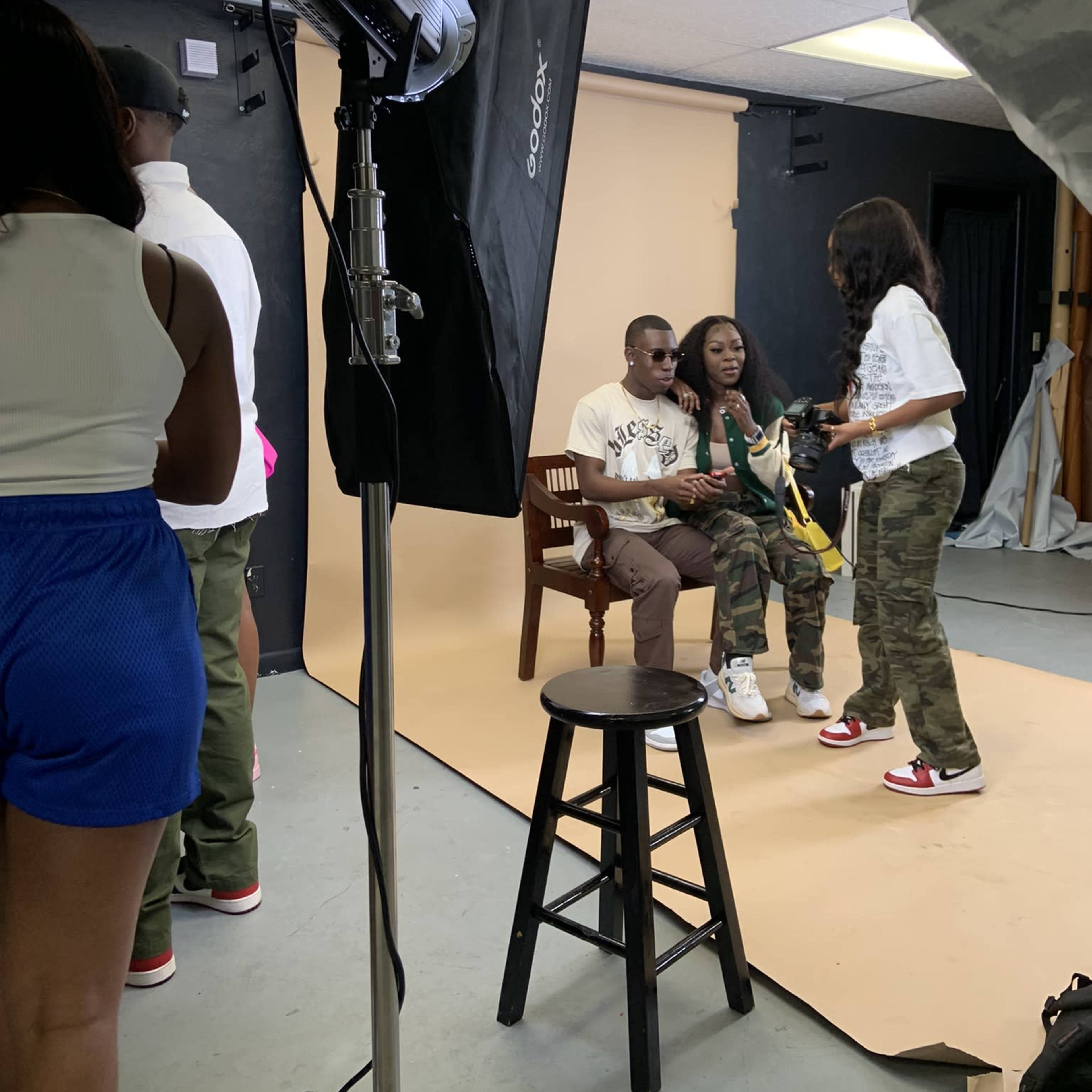 A photoshoot takes place in a studio with subjects posing on a chair against a backdrop, while a photographer and assistants manage lighting and setup.